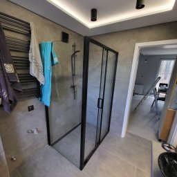 Nowi-Rem Paweł Nowicki - Nowoczesna kabina prysznicowa z czarnymi profilami, szare płytki, grzejnik drabinkowy z ręcznikami, widok na remontowane pomieszczenie z drabiną.