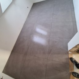 Nowi-Rem Paweł Nowicki - Ułożone szare płytki podłogowe w korytarzu, widoczne ślady po fugowaniu, fragment obuwia w prawym dolnym rogu.