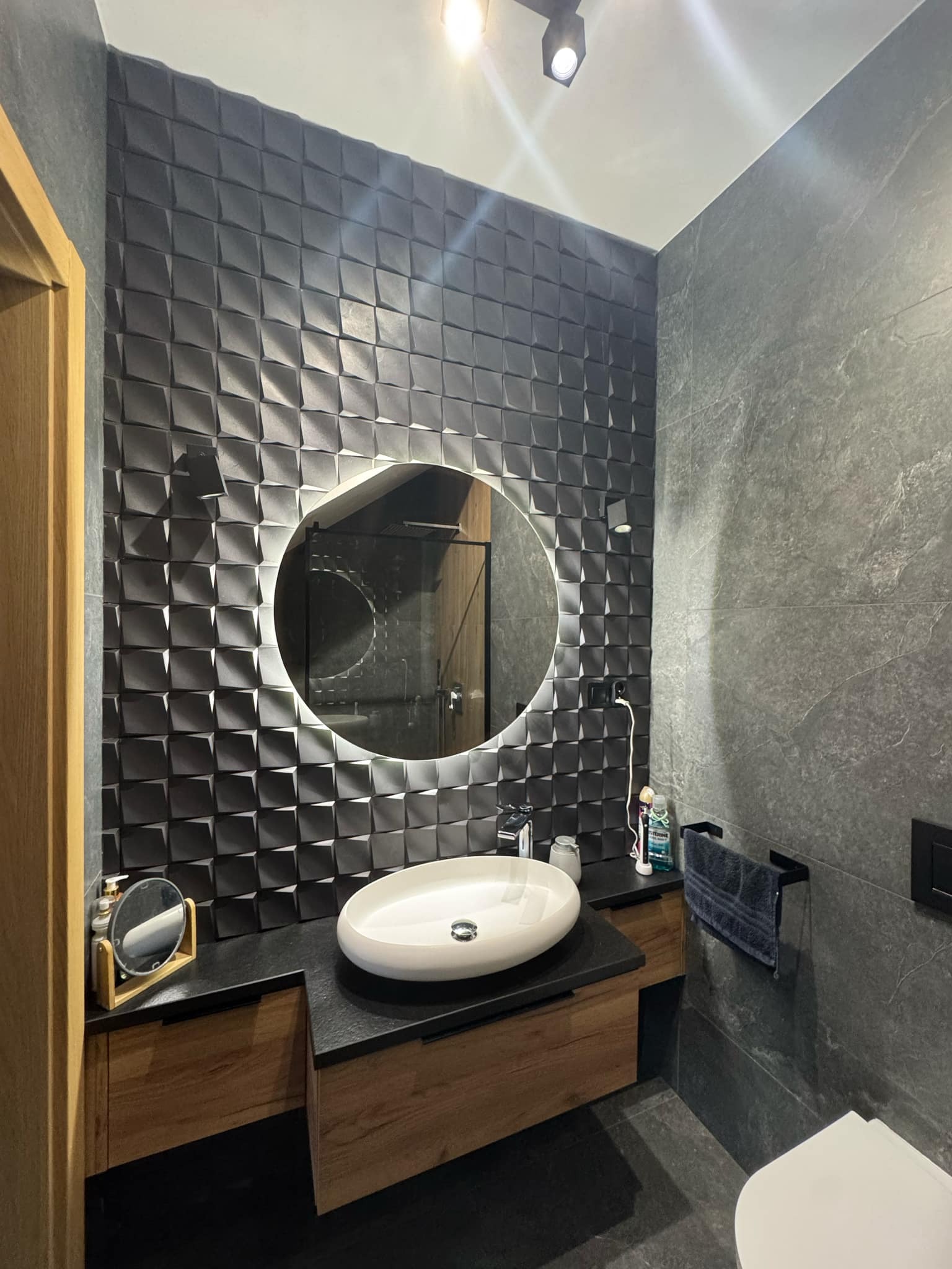 Nowoczesna łazienka z czarną, trójwymiarową ścianą, owalną umywalką nablatową i okrągłym lustrem z podświetleniem LED.