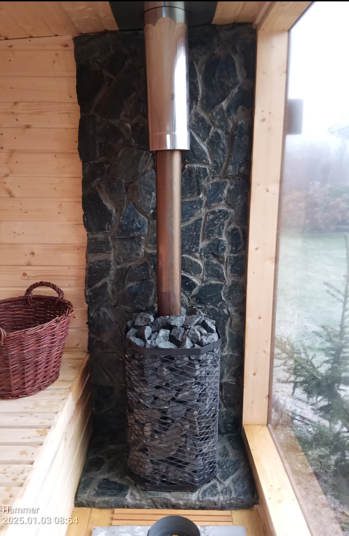 Wnętrze sauny z piecem opalanym drewnem obłożonym kamieniami, komin z metalu, ściana wyłożona kamieniem, drewniane ławki i koszyk wiklinowy.