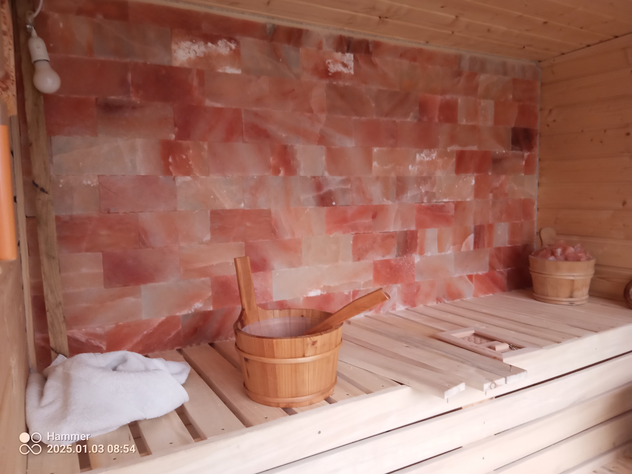 Wnętrze sauny z drewnianymi ławami, ścianą z bloków soli himalajskiej, drewnianym wiadrem z chochlą i miską z solą na ławie. Oświetlenie punktowe podkreśla fakturę soli.