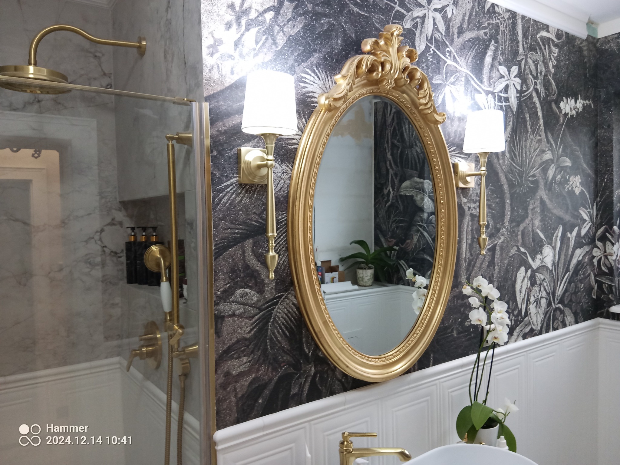 Elegancka łazienka z motywem roślinnym na ścianie, złota armatura, owalne lustro w ozdobnej ramie i białe orchidee.