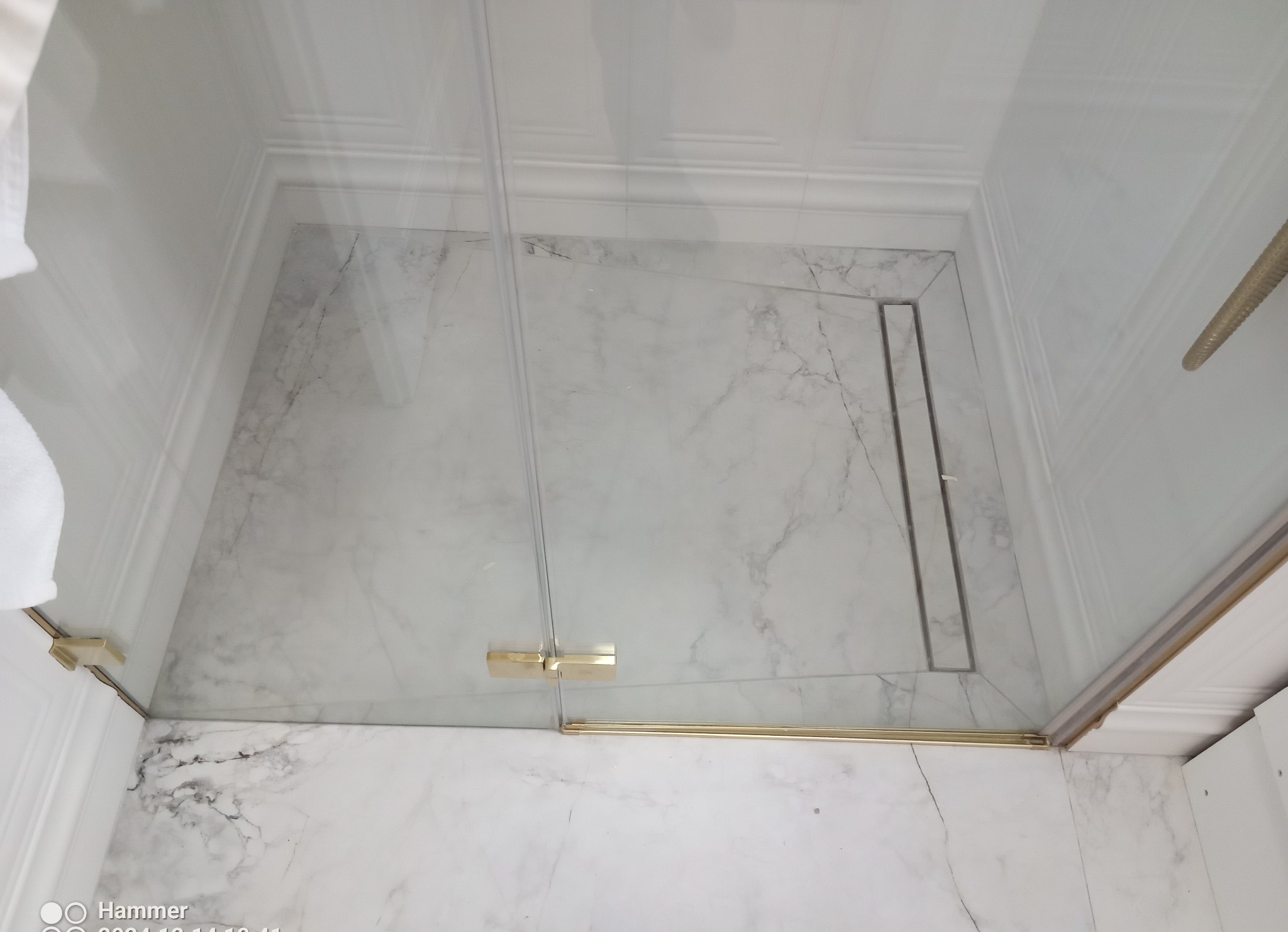 Nowoczesna kabina prysznicowa walk-in z odpływem liniowym, wykończona płytkami imitującymi biały marmur z delikatnym użyleniem, złote detale wykończeniowe.