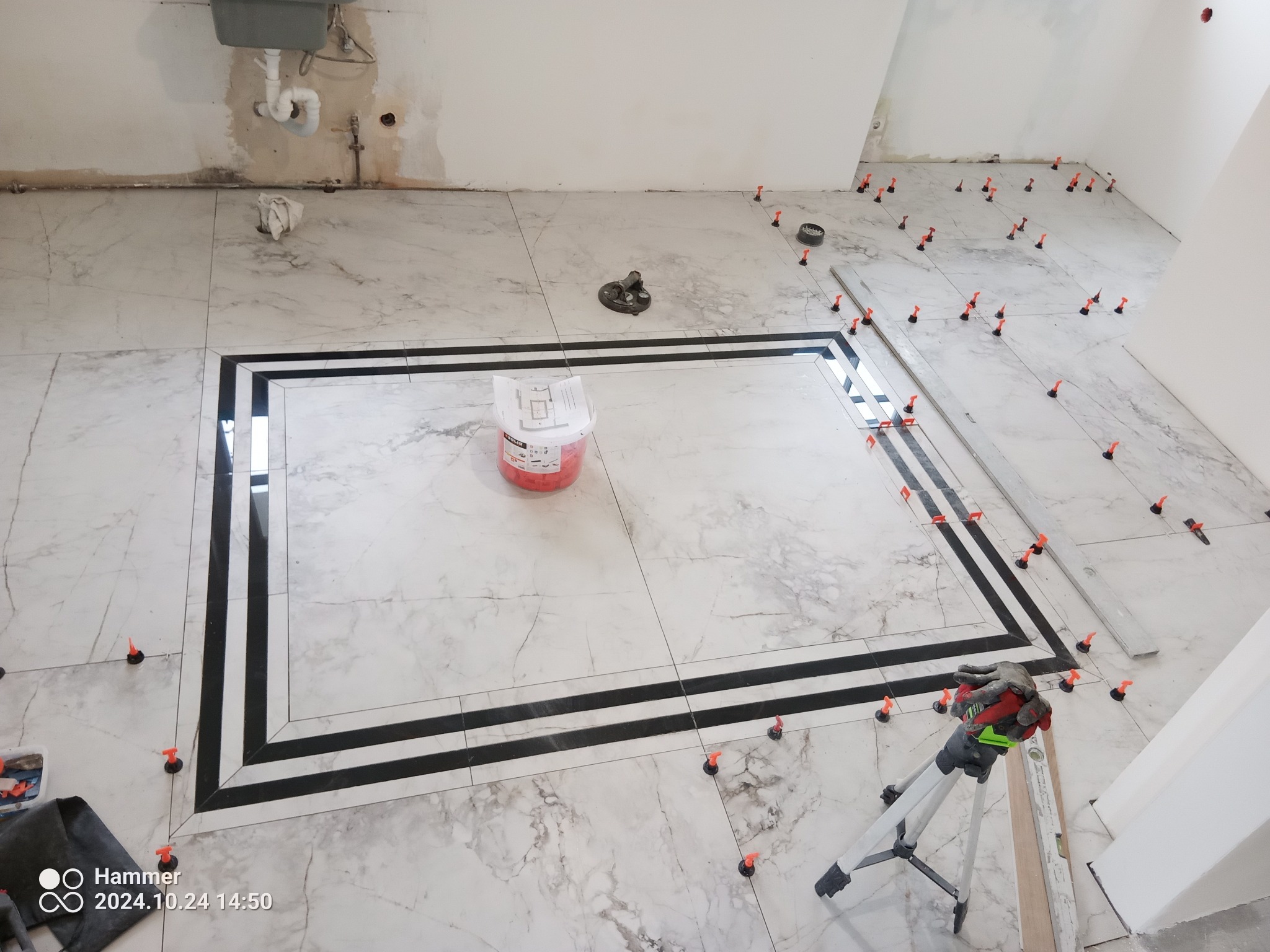 Podłoga w trakcie układania płytek marmuropodobnych z czarną bordiurą, widoczne narzędzia glazurnicze: poziomica, przyssawka do płytek, wiadro z zaprawą i system poziomowania płytek.