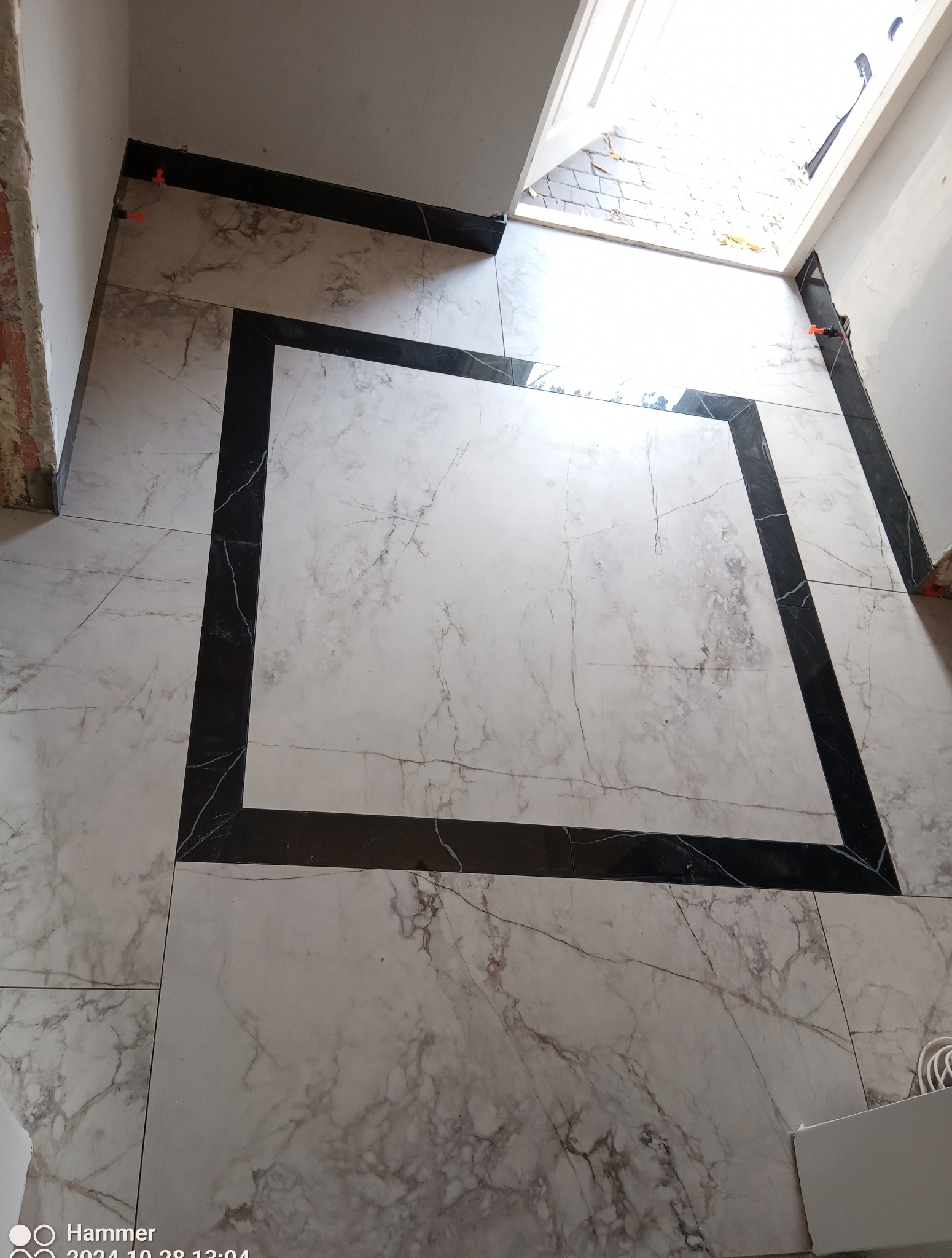 Ułożona podłoga z płytek imitujących marmur, obramowana czarnymi płytkami, widoczne pomarańczowe kliny dystansowe, surowy stan wykończenia ściany i widok na zewnątrz przez niedokończone drzwi.