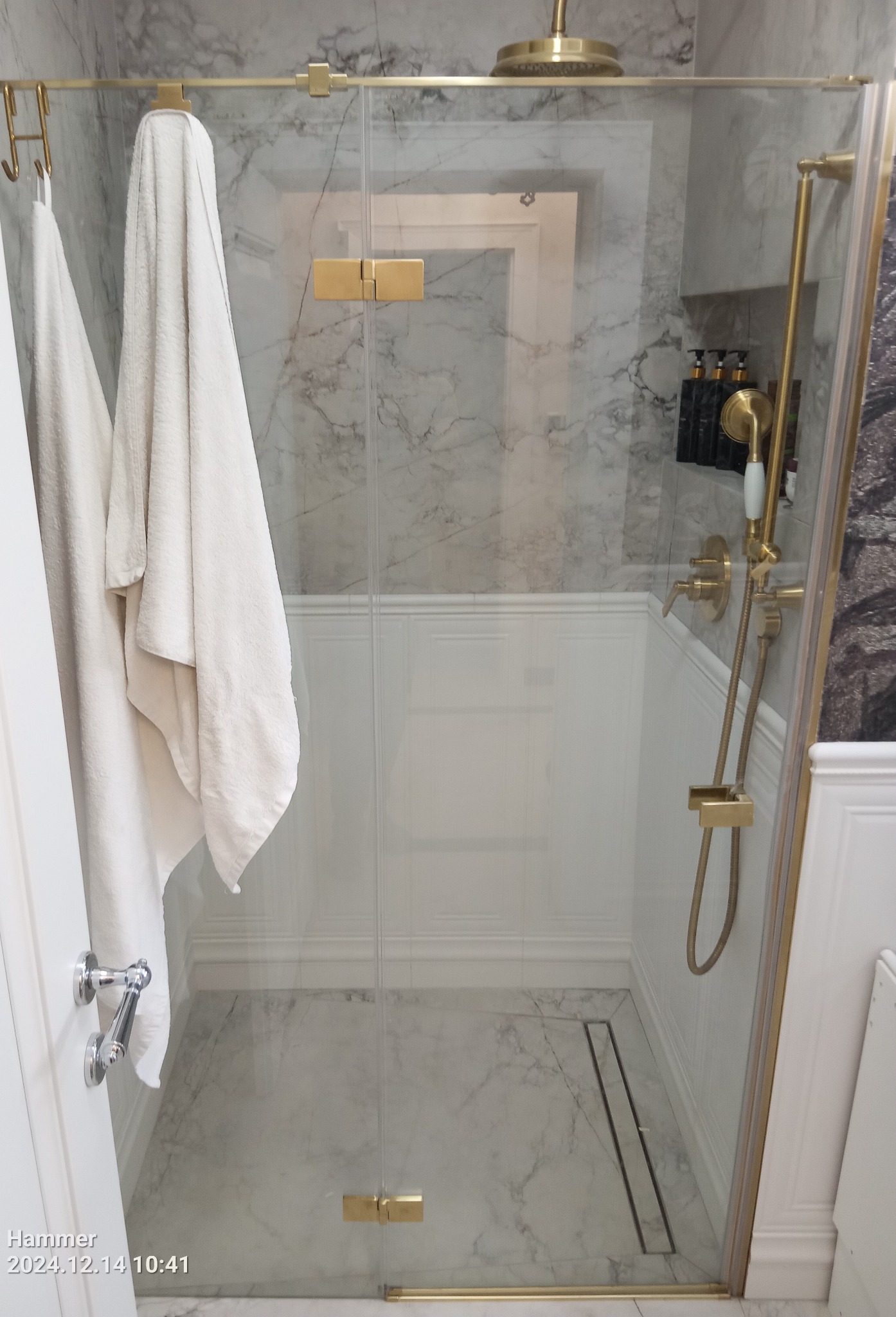 Szklana kabina prysznicowa z złotymi okuciami, marmurowe płytki, białe ręczniki na haczykach, deszczownica i odpływ liniowy.