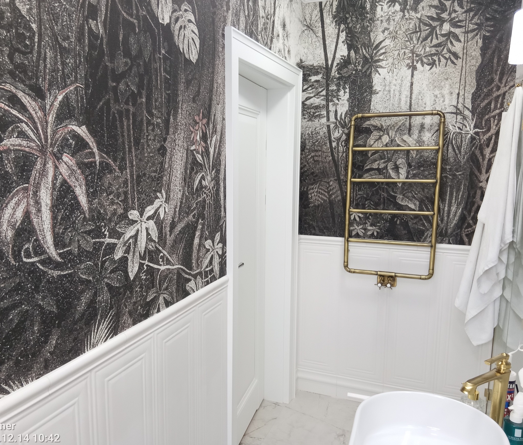 Nowoczesna łazienka z tapetą w motywy roślinne, białą boazerią i złotym grzejnikiem drabinkowym, widok na umywalkę i fragment lustra.