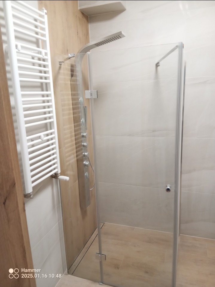 Nowoczesna kabina prysznicowa ze szklanymi drzwiami, panel prysznicowy z hydromasażem oraz biały grzejnik drabinkowy w wykończonej łazience z płytkami imitującymi drewno.