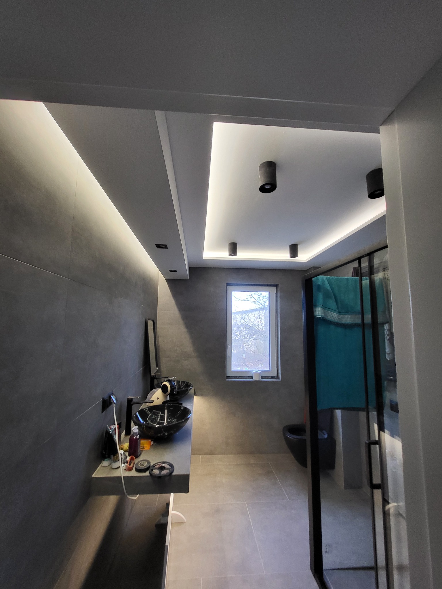 Nowoczesna łazienka z szarymi płytkami, podwieszanymi umywalkami w kolorze czarnym z białymi żyłkami, oświetleniem LED podsufitowym i kabiną prysznicową z czarnymi ramami.