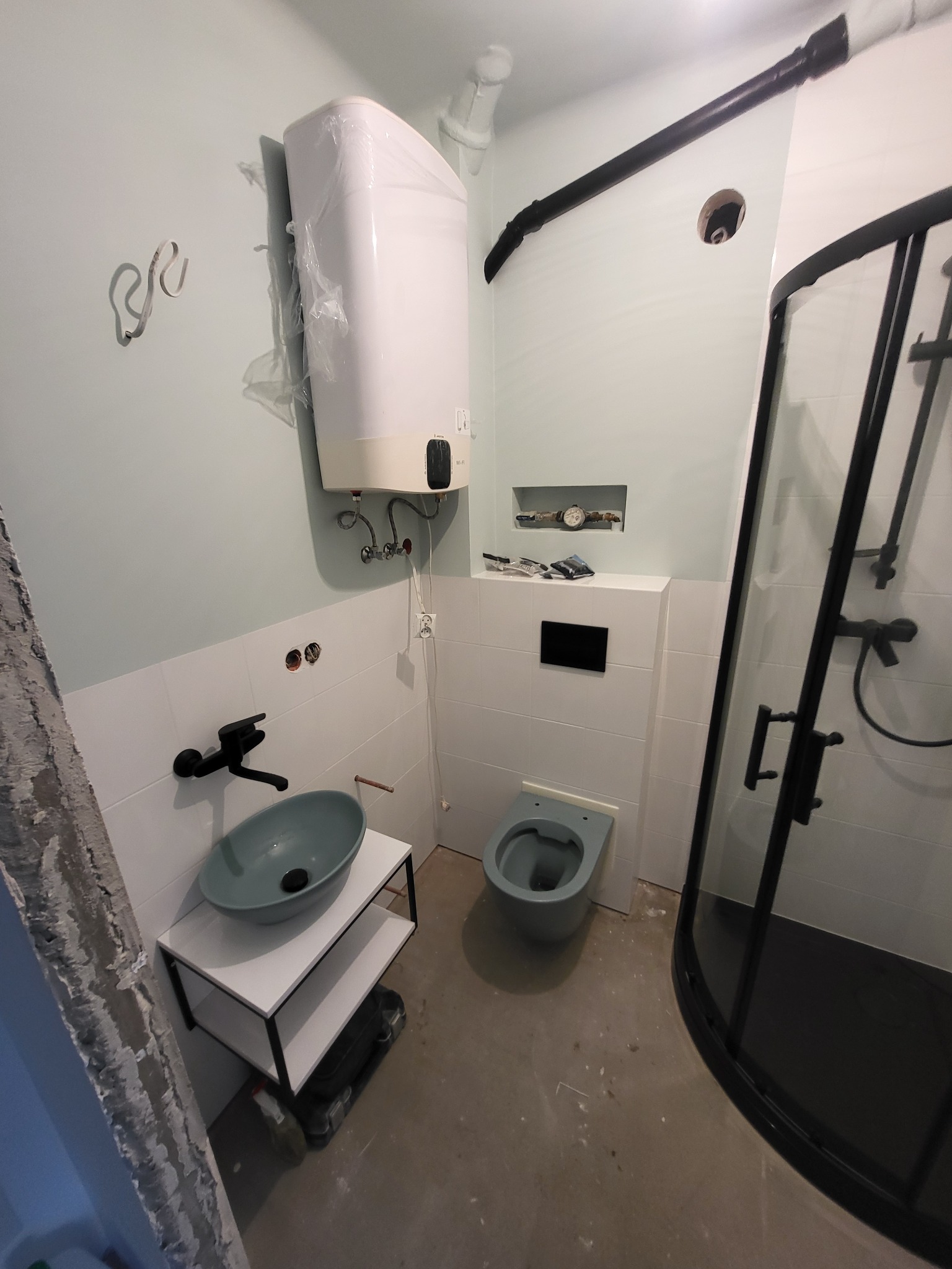 Niedokończona łazienka z zamontowanym bojlerem w folii, umywalką na stelażu, toaletą i kabiną prysznicową z czarnymi ramami. Widoczne narzędzia i niedociągnięcia wykończeniowe.