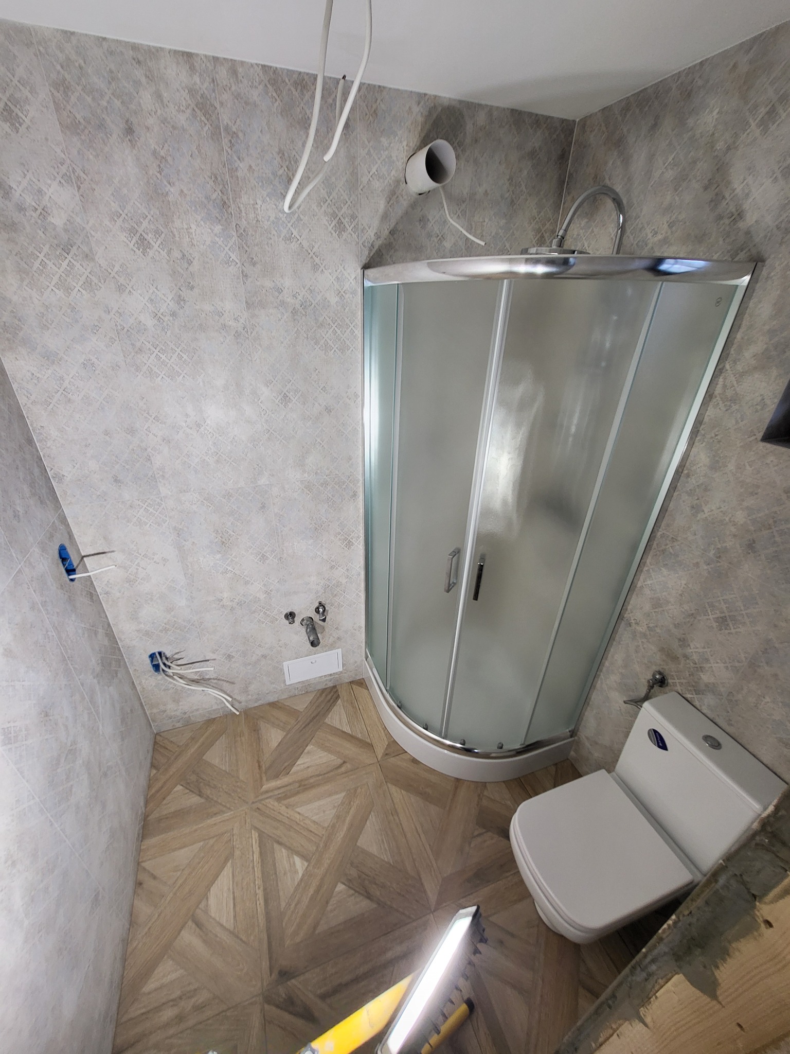 Narożna kabina prysznicowa z matowego szkła, toaleta i drewnopodobne płytki podłogowe w trakcie remontu łazienki; widoczne wystające kable elektryczne i rury doprowadzające wodę.