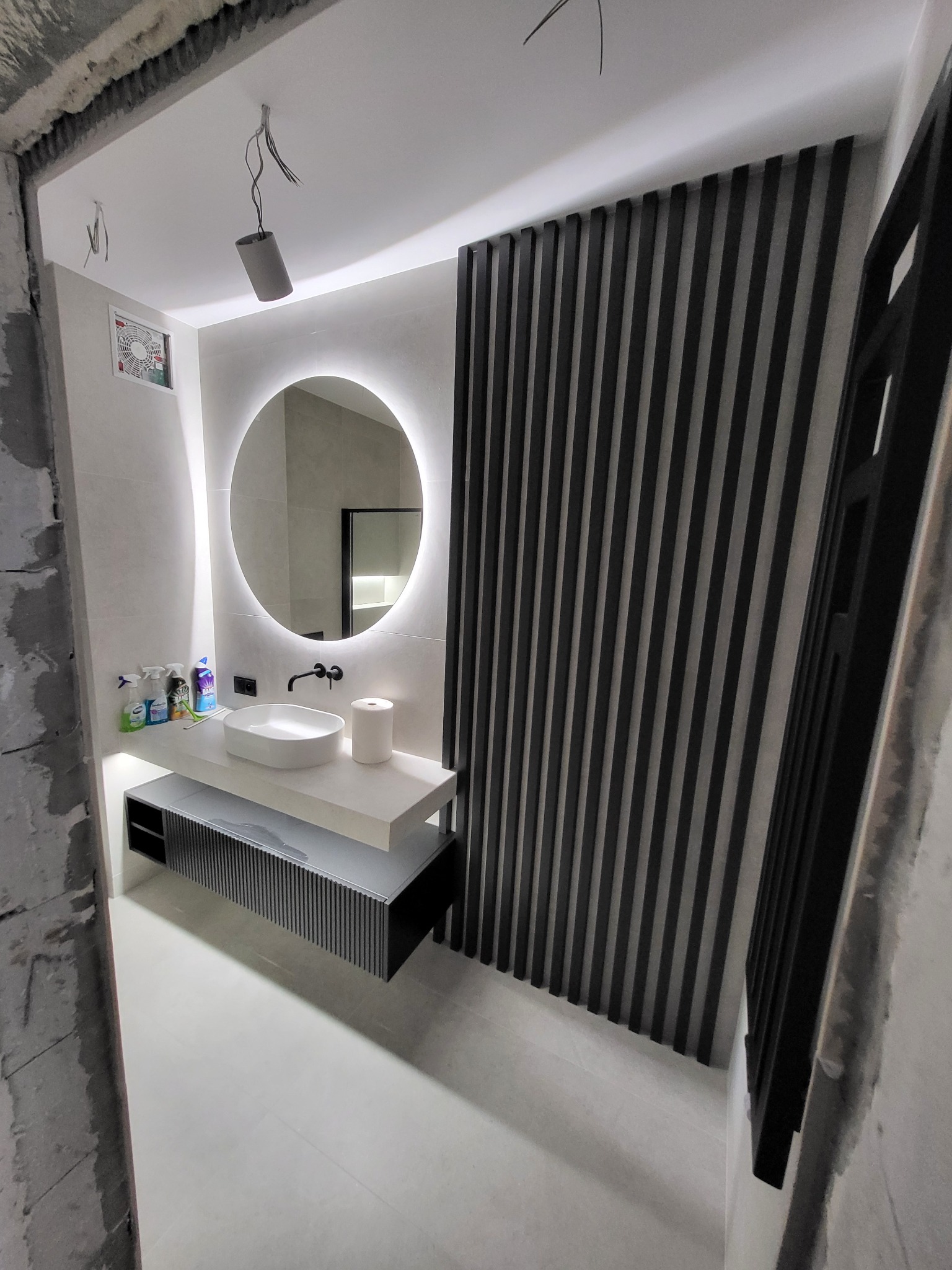 Nowoczesna łazienka w trakcie remontu: białe płytki, okrągłe lustro z podświetleniem LED, umywalka nablatowa, czarna armatura i dekoracyjna ścianka z pionowych, czarnych listew.