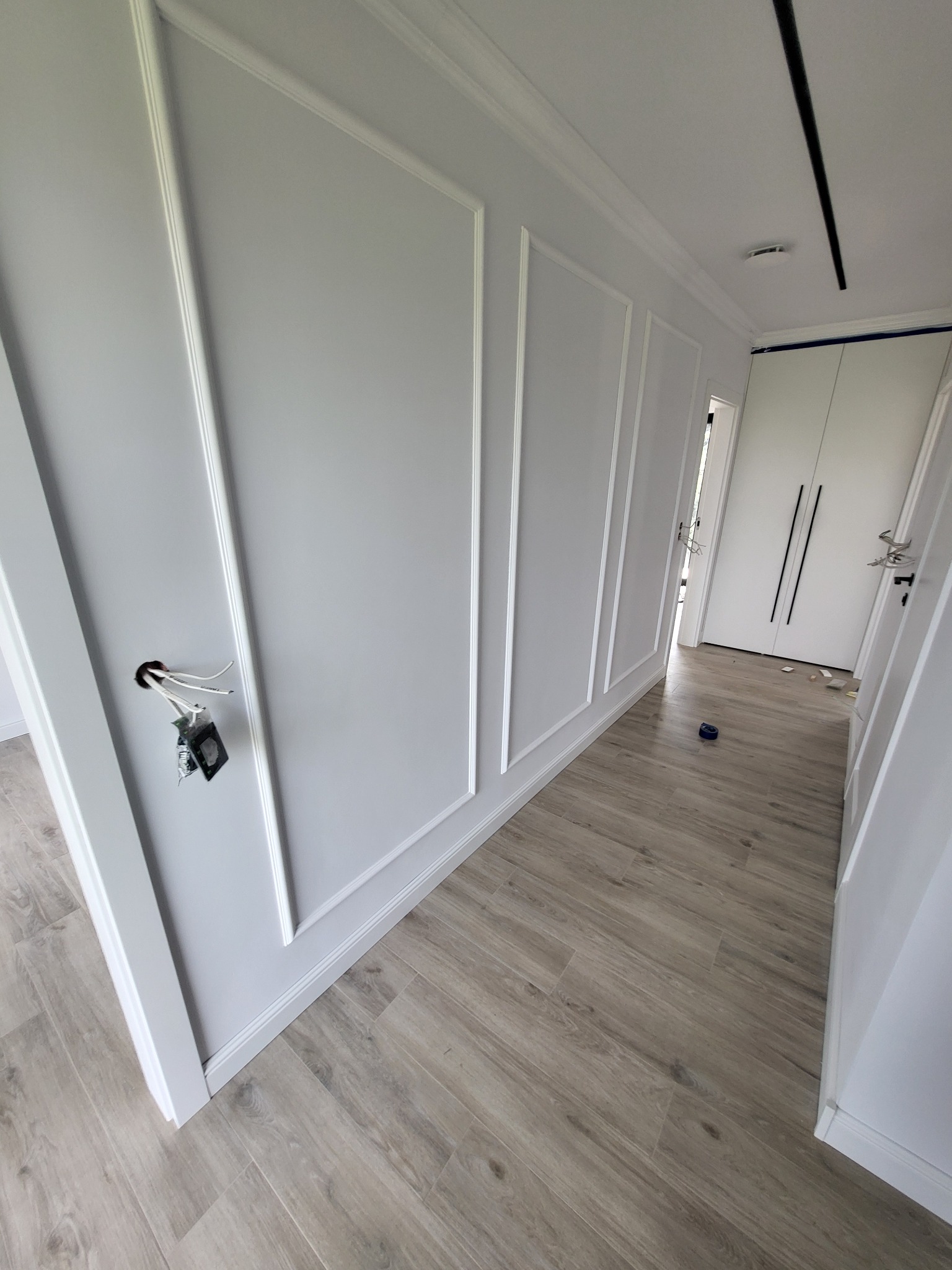 Korytarz w trakcie wykańczania: szare ściany z białymi listwami dekoracyjnymi, drewnopodobne panele na podłodze, wystające kable ze ściany w miejscu gniazdka elektrycznego.