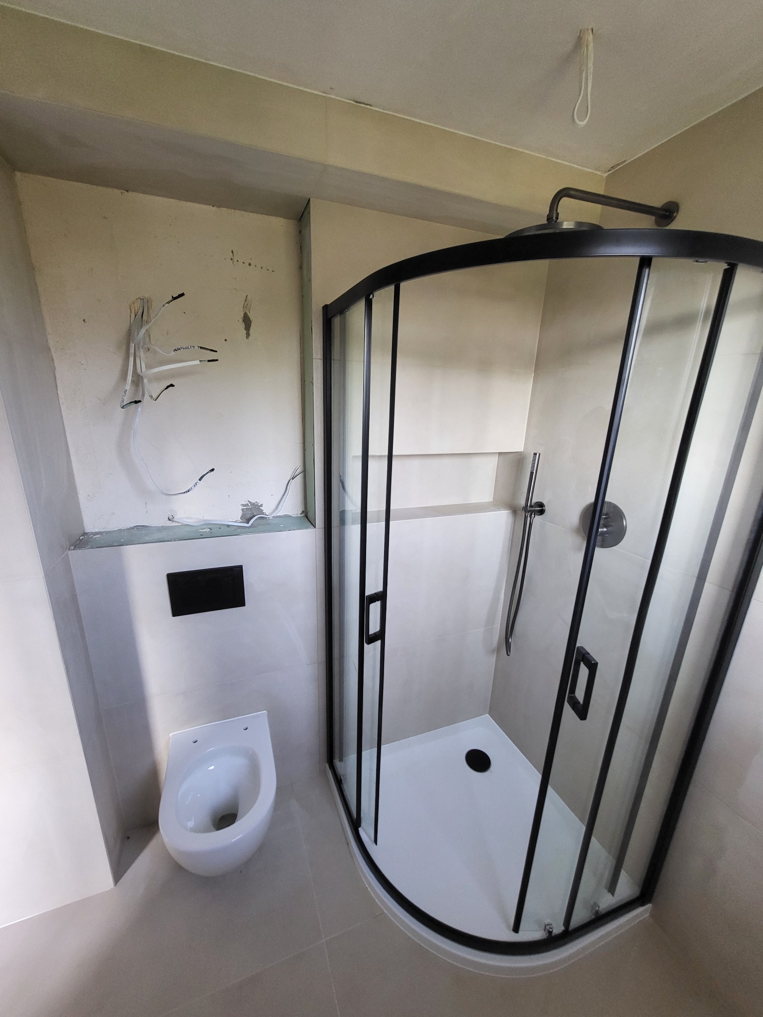 Niedokończona łazienka z kabiną prysznicową z czarnymi ramami, wiszącą toaletą i wystającymi przewodami elektrycznymi ze ściany.