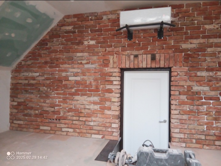 Ściana z cegły klinkierowej z zamontowanym drążkiem do podciągania i białymi drzwiami, widoczne narzędzia budowlane na podłodze, ściana częściowo zagruntowana.