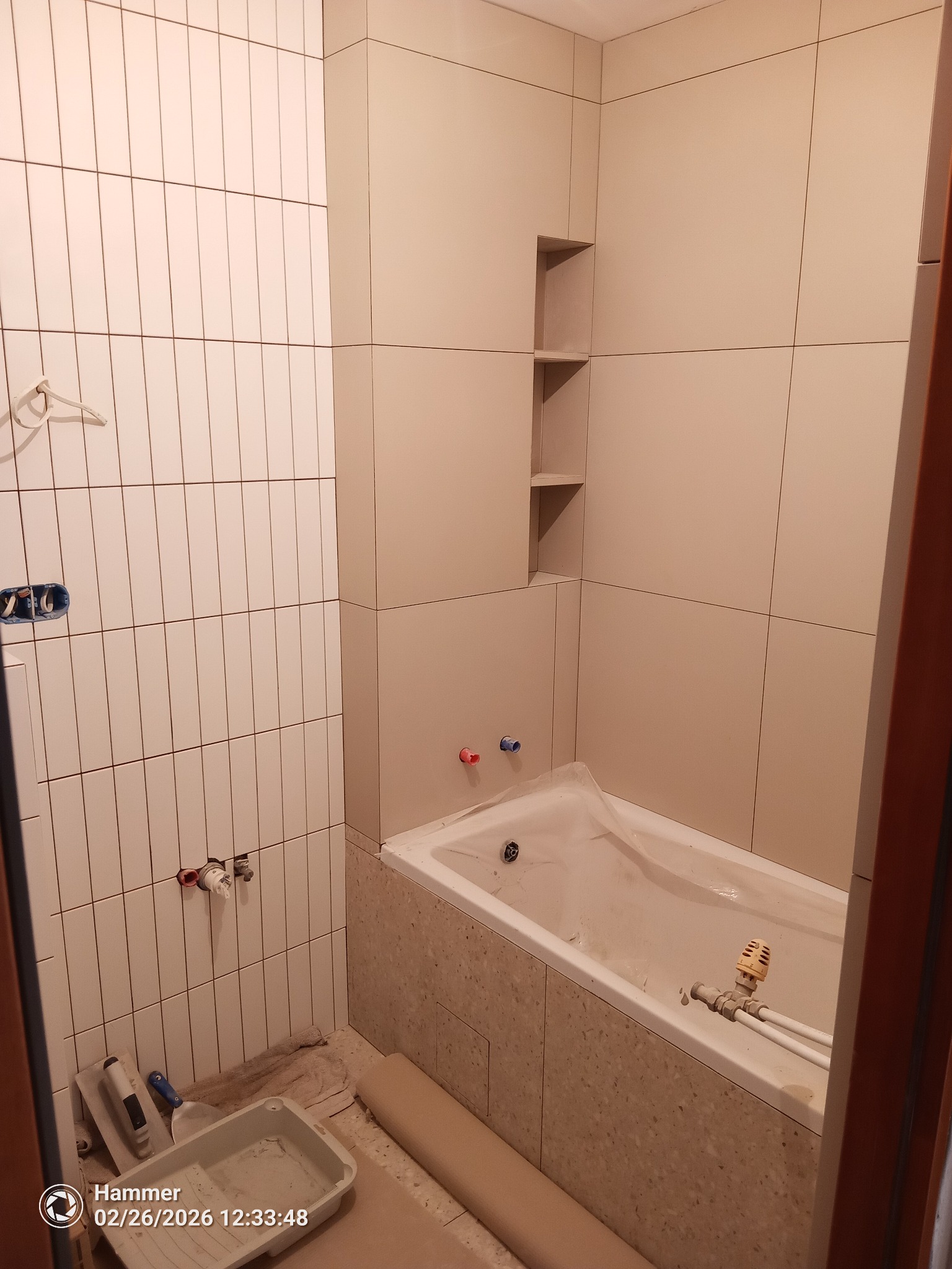 Jasna łazienka w trakcie prac: beżowe płytki ścienne ułożone w pionie i poziomie, wnęka z półkami, wanna zabezpieczona folią, widoczne narzędzia glazurnicze.