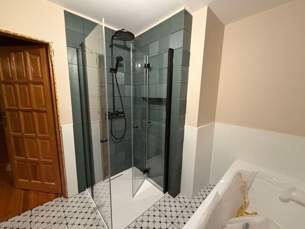 Nowoczesna kabina prysznicowa z czarnymi elementami, zielone płytki strukturalne, wzorzysta podłoga i drewniane drzwi w tle. Widok na fragment wanny.