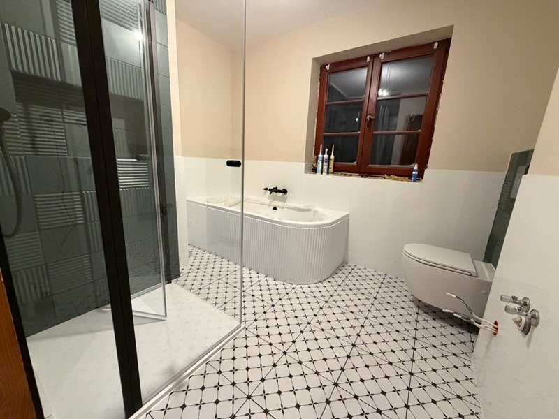 Nowoczesna łazienka po remoncie w Niedziałce Drugiej: kabina prysznicowa z czarną ramą, biała wanna z żłobieniami, toaleta i wzorzysta podłoga. Okno z drewnianą ramą.