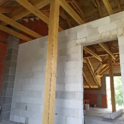 Wnętrze w trakcie budowy z widocznymi ścianami z białych bloczków, drewnianą konstrukcją dachu i słupem podtrzymującym, z fragmentami ścian z czerwonej cegły w tle.