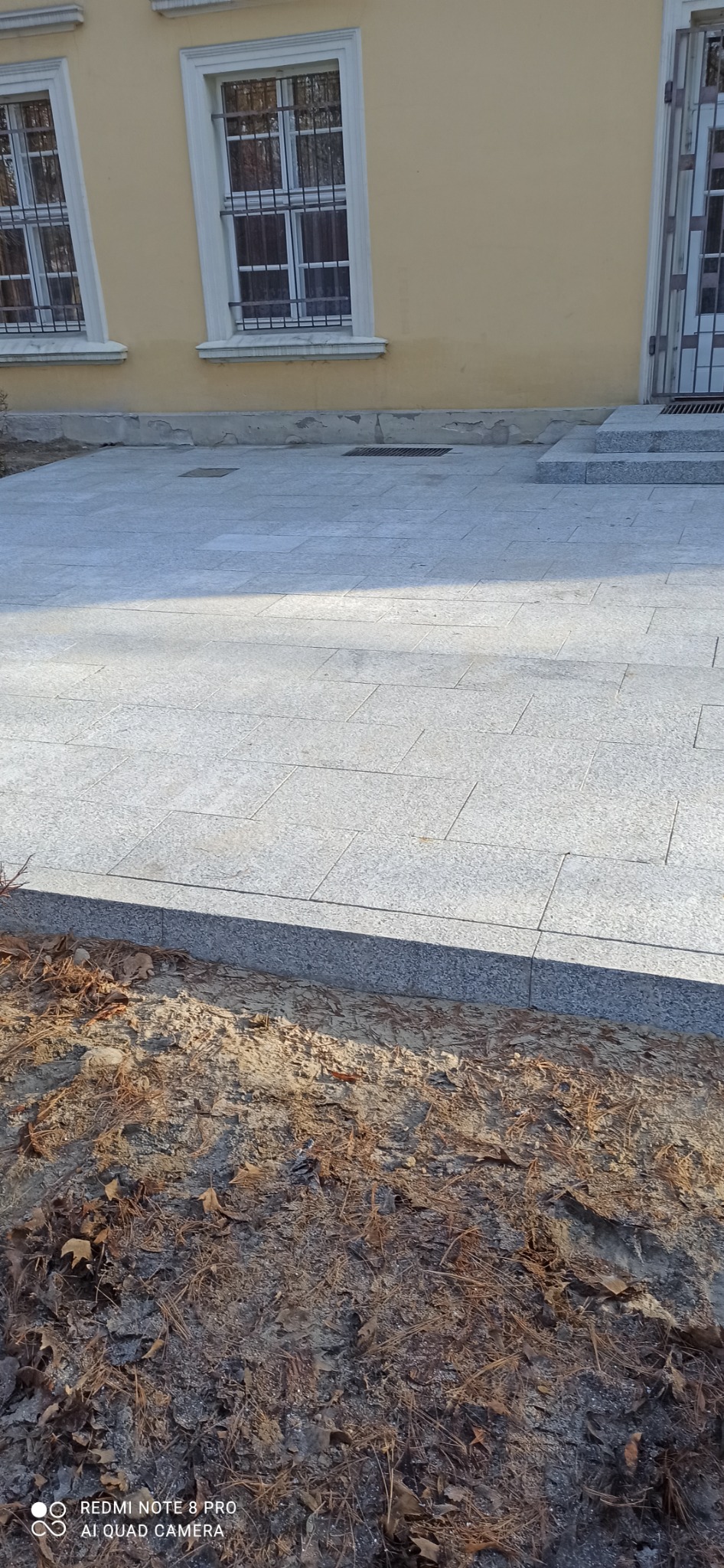 Wyłożony jasnoszarą kostką brukową chodnik przed budynkiem z żółtą elewacją, widoczne okna z kratami i schody wejściowe.