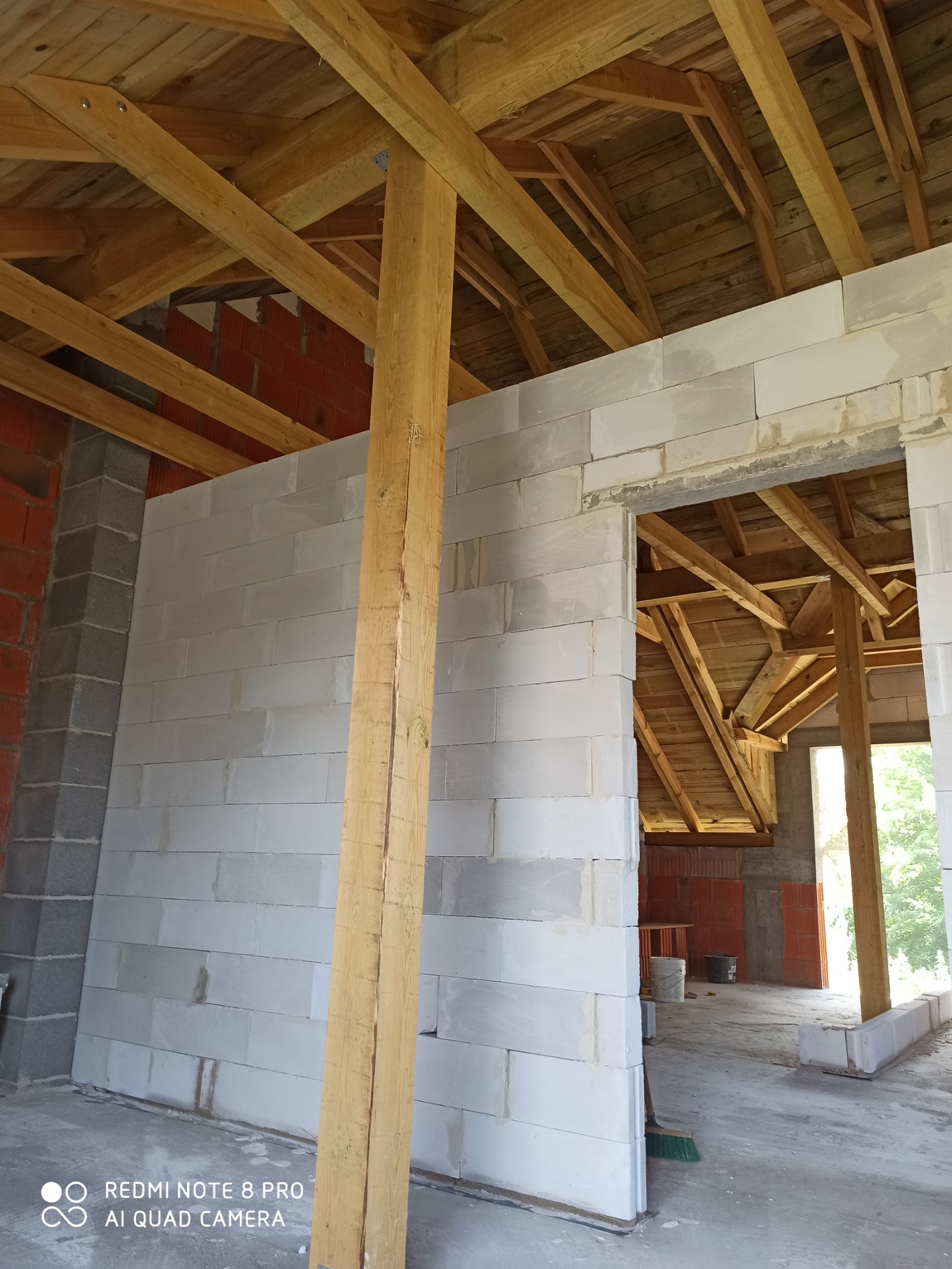 Wnętrze w trakcie budowy z widocznymi ścianami z białych bloczków, drewnianą konstrukcją dachu i słupem podtrzymującym, z fragmentami ścian z czerwonej cegły w tle.