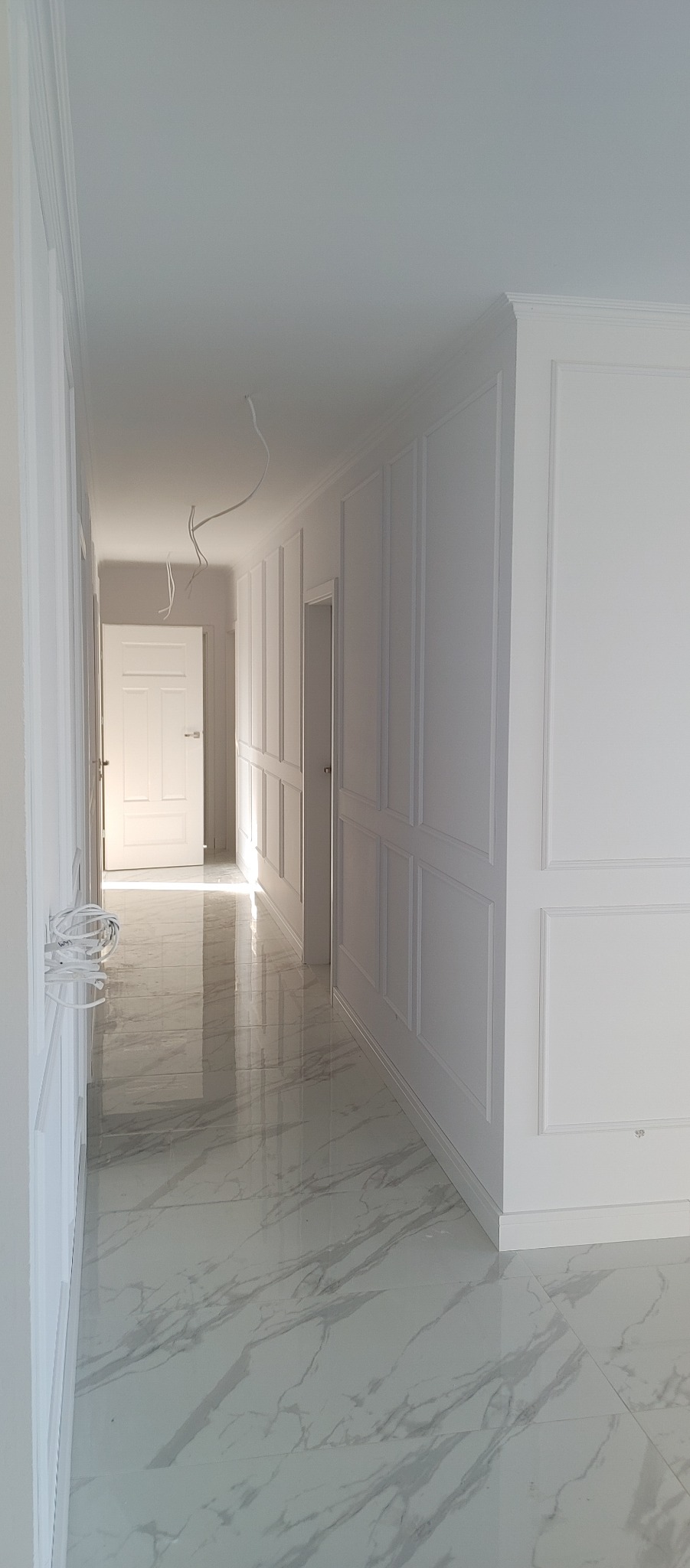 Biały korytarz z panelami ściennymi i marmurową podłogą, widoczne wiszące przewody elektryczne.