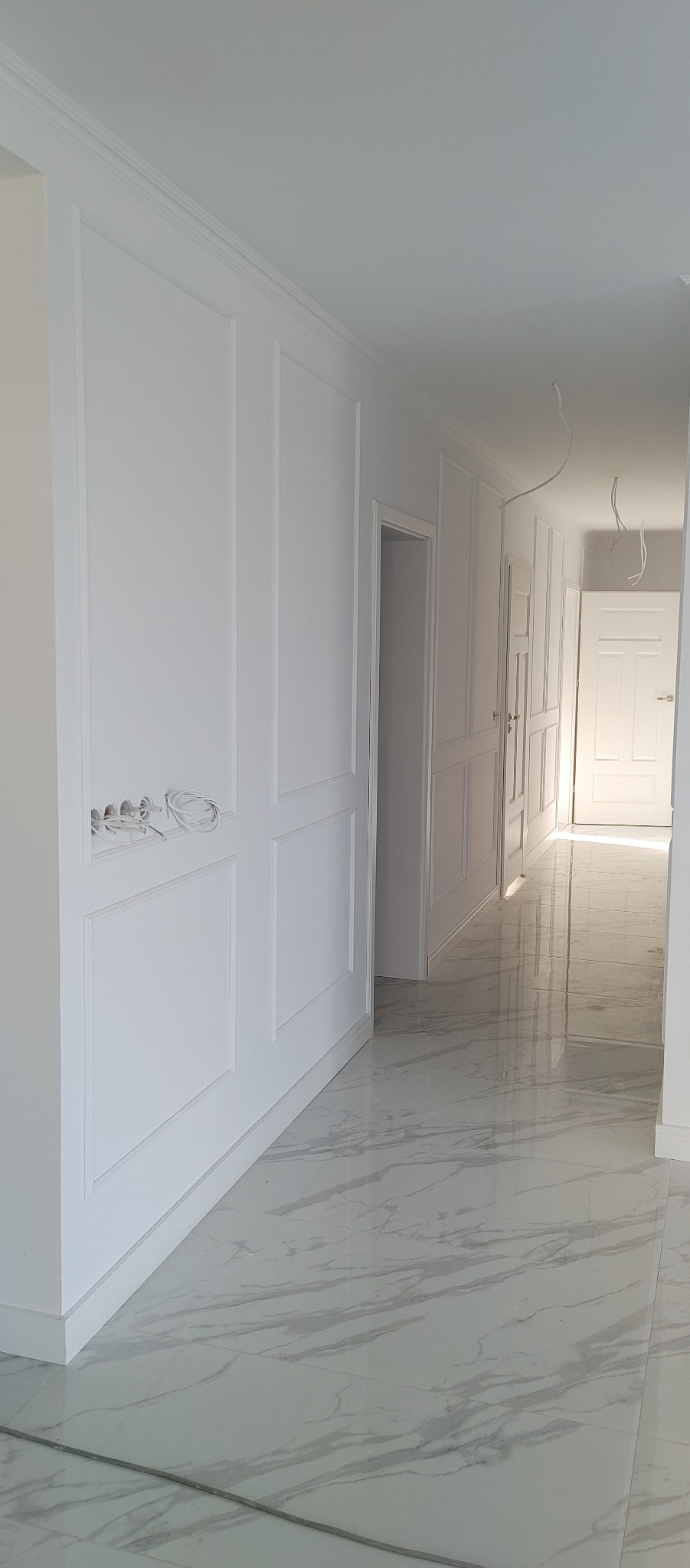 Biały korytarz z panelami ściennymi i marmurową podłogą w odcieniach szarości, widoczne wystające kable elektryczne z puszki w ścianie.