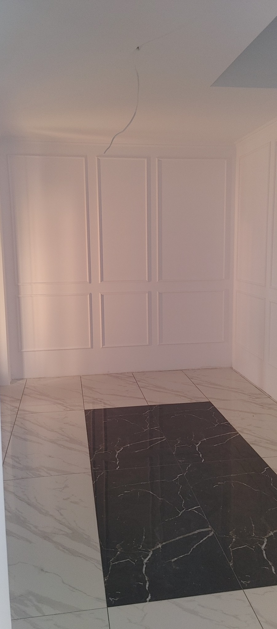 Wykończone wnętrze z panelami ściennymi i kontrastującą podłogą z płytek marmurowych w odcieniach bieli i czerni. Na suficie widoczny kabel instalacji elektrycznej.