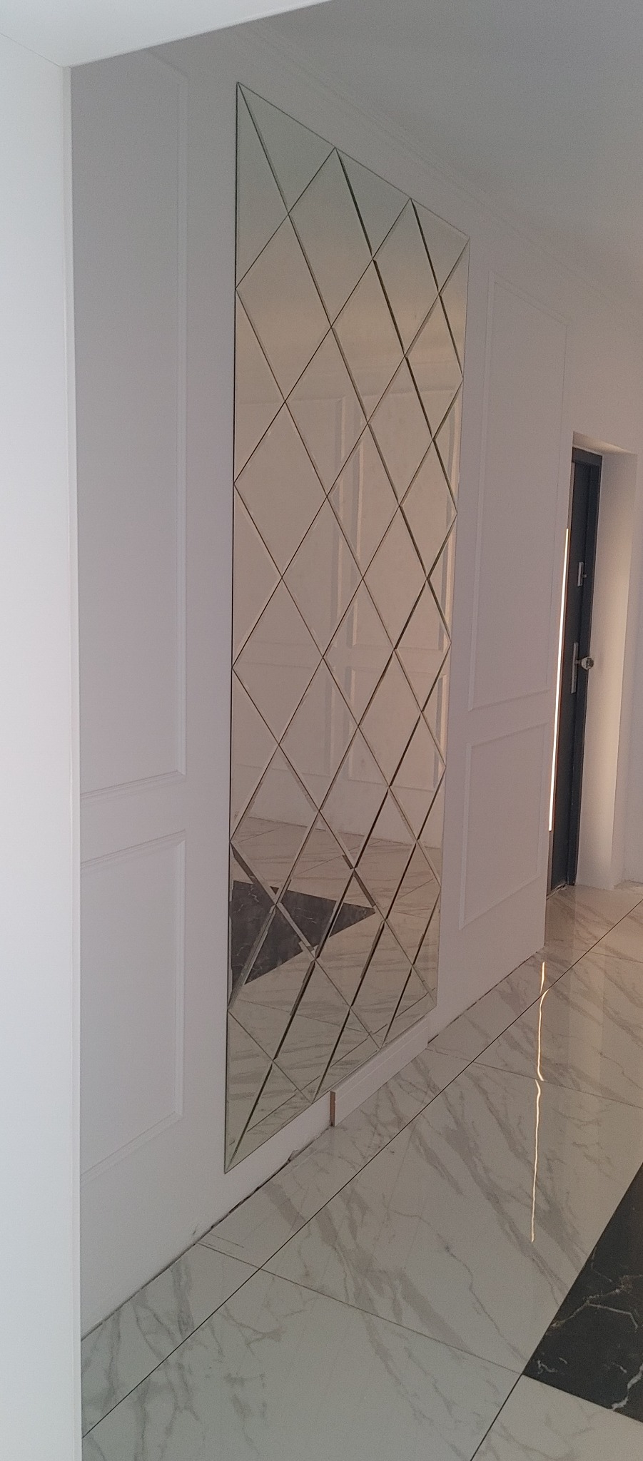 Ściana w korytarzu z dekoracyjnym lustrem złożonym z romboidalnych płytek, odbijających marmurową podłogę i fragment drzwi.