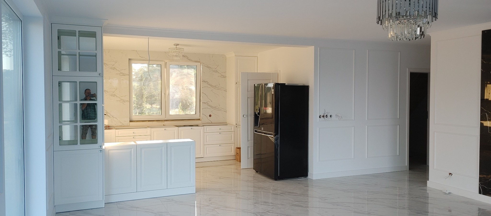Nowoczesna kuchnia z białymi szafkami, marmurowymi płytkami i czarną lodówką; widoczne niedokończone podłączenia elektryczne i hydrauliczne.