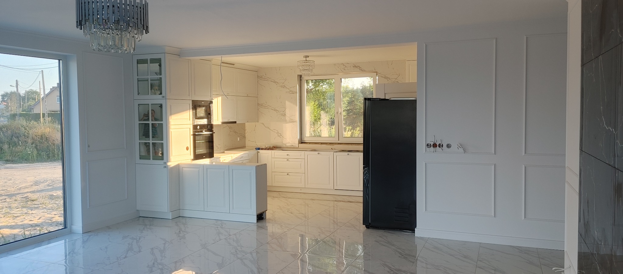 Nowoczesna kuchnia w stylu klasycznym, białe meble z frezowanymi frontami, marmurowe płytki podłogowe z połyskiem, czarna lodówka, ozdobne listwy ścienne, widoczne gniazdka elektryczne przed...