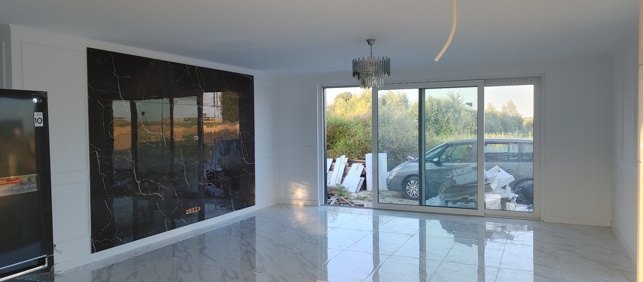 Wykończone wnętrze: czarny marmur na ścianie, błyszcząca podłoga z płytek imitujących marmur, przesuwne drzwi balkonowe, widoczne materiały budowlane na zewnątrz.