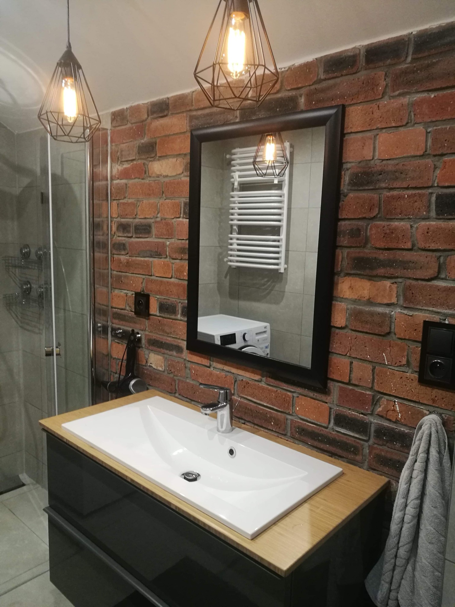 Nowoczesna łazienka z umywalką nablatową, szafką w kolorze grafitowym z drewnianym blatem, ściana z czerwonej cegły, lustro w czarnej ramie, lampy wiszące w stylu industrialnym i kabina prysznicowa...