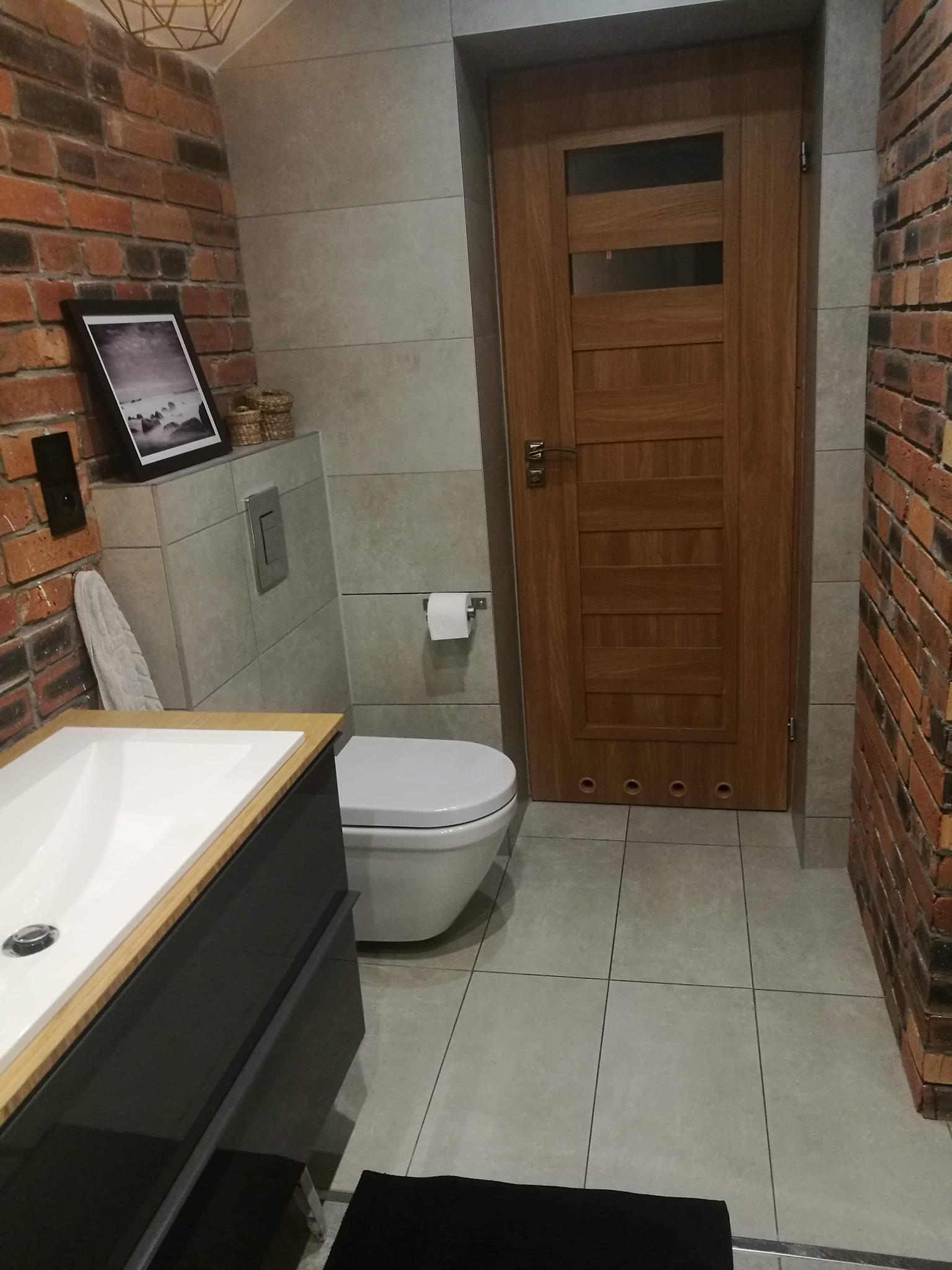 Wykończona łazienka z ceglaną ścianą, umywalką z szafką w kolorze grafitowym, toaletą i drewnianymi drzwiami z przeszkleniami, oświetlona geometryczną lampą sufitową.