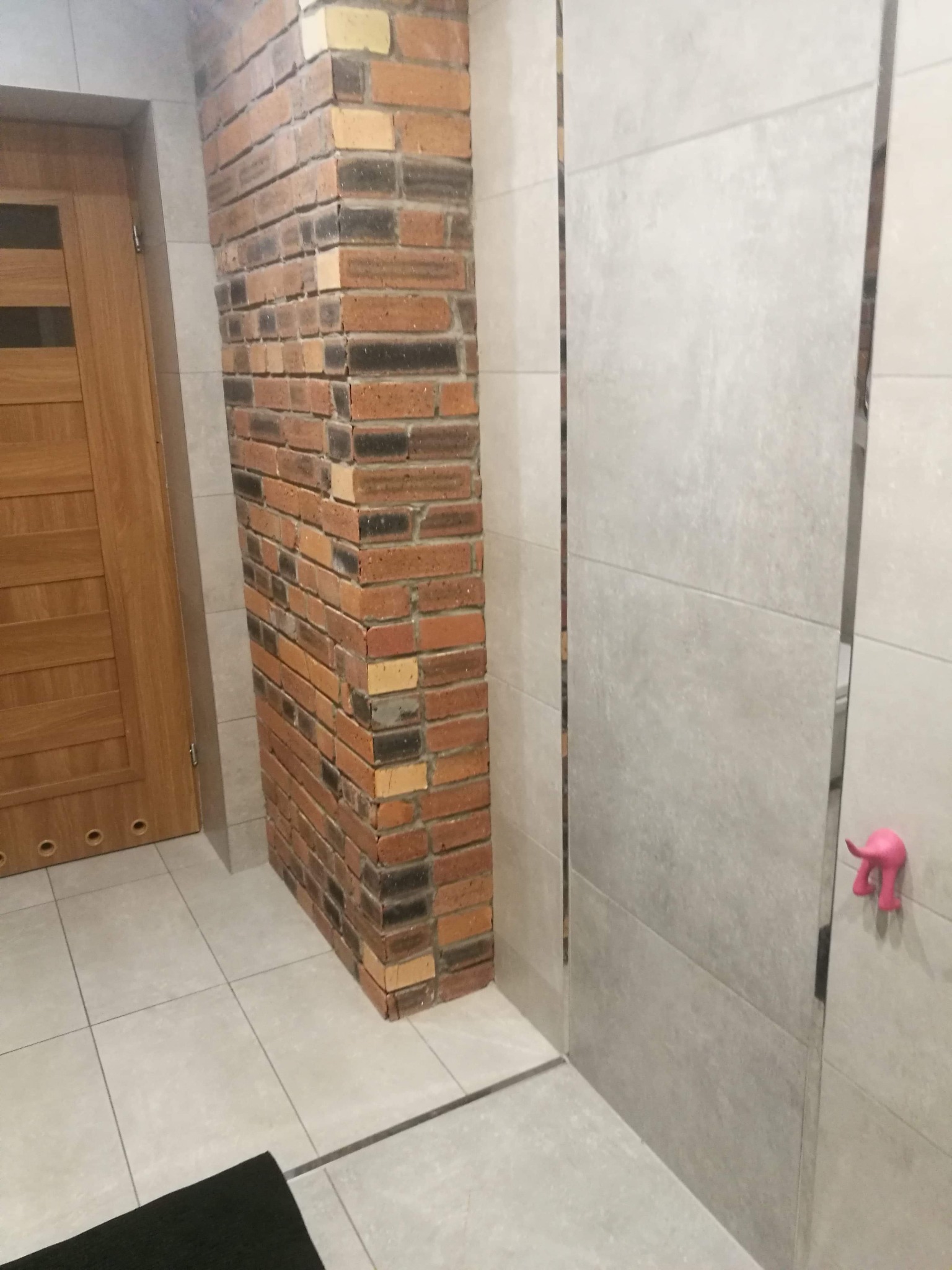 Wykończona łazienka z ceglanym filarem, drewnianymi drzwiami i szarą okładziną ścienną z różowym wieszakiem w kształcie słonia.