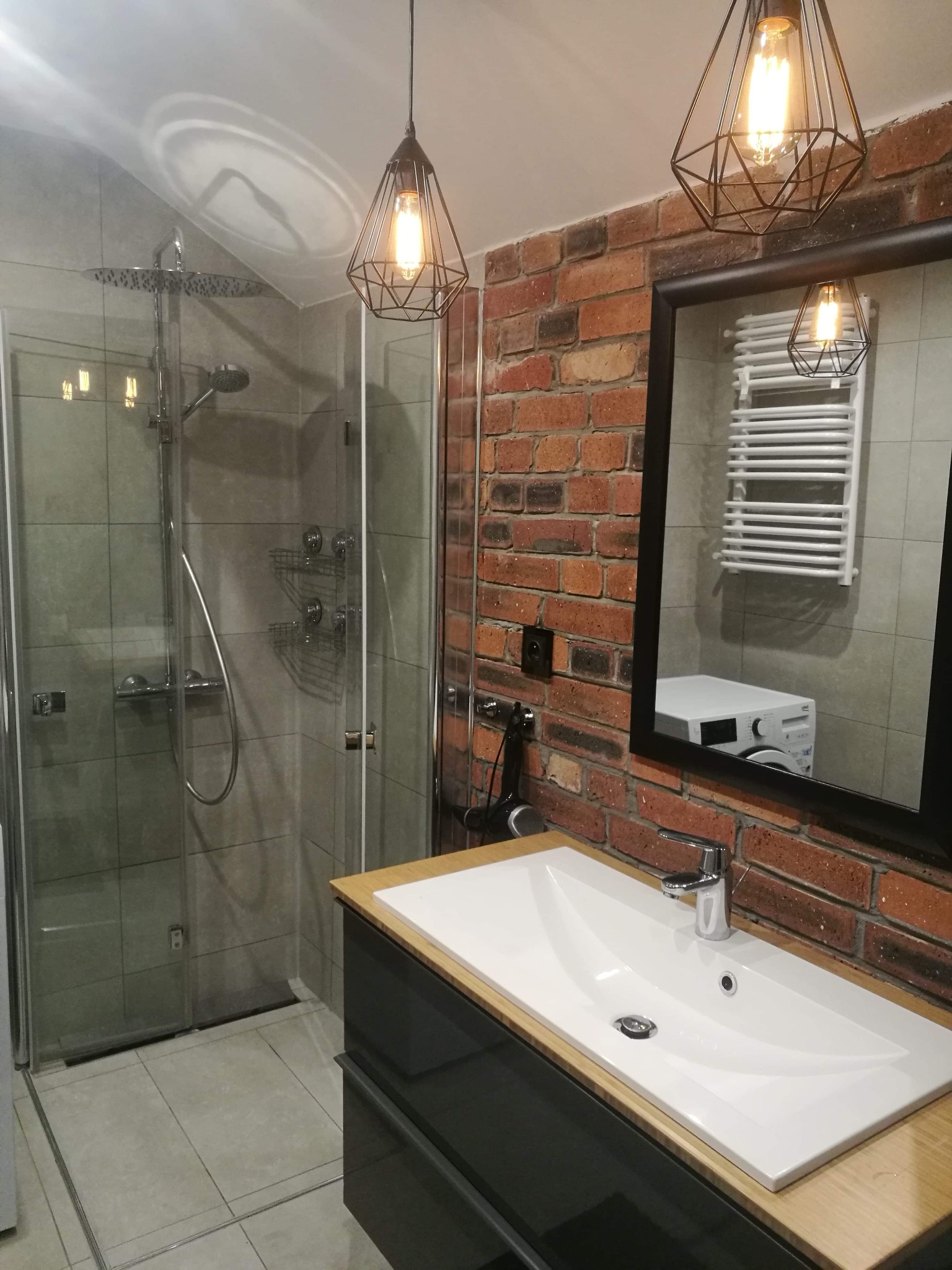 Nowoczesna łazienka z kabiną prysznicową, umywalką nablatową i ścianą z cegły, oświetlona lampami wiszącymi w stylu industrialnym, z widocznym odbiciem grzejnika drabinkowego i pralki w lustrze.