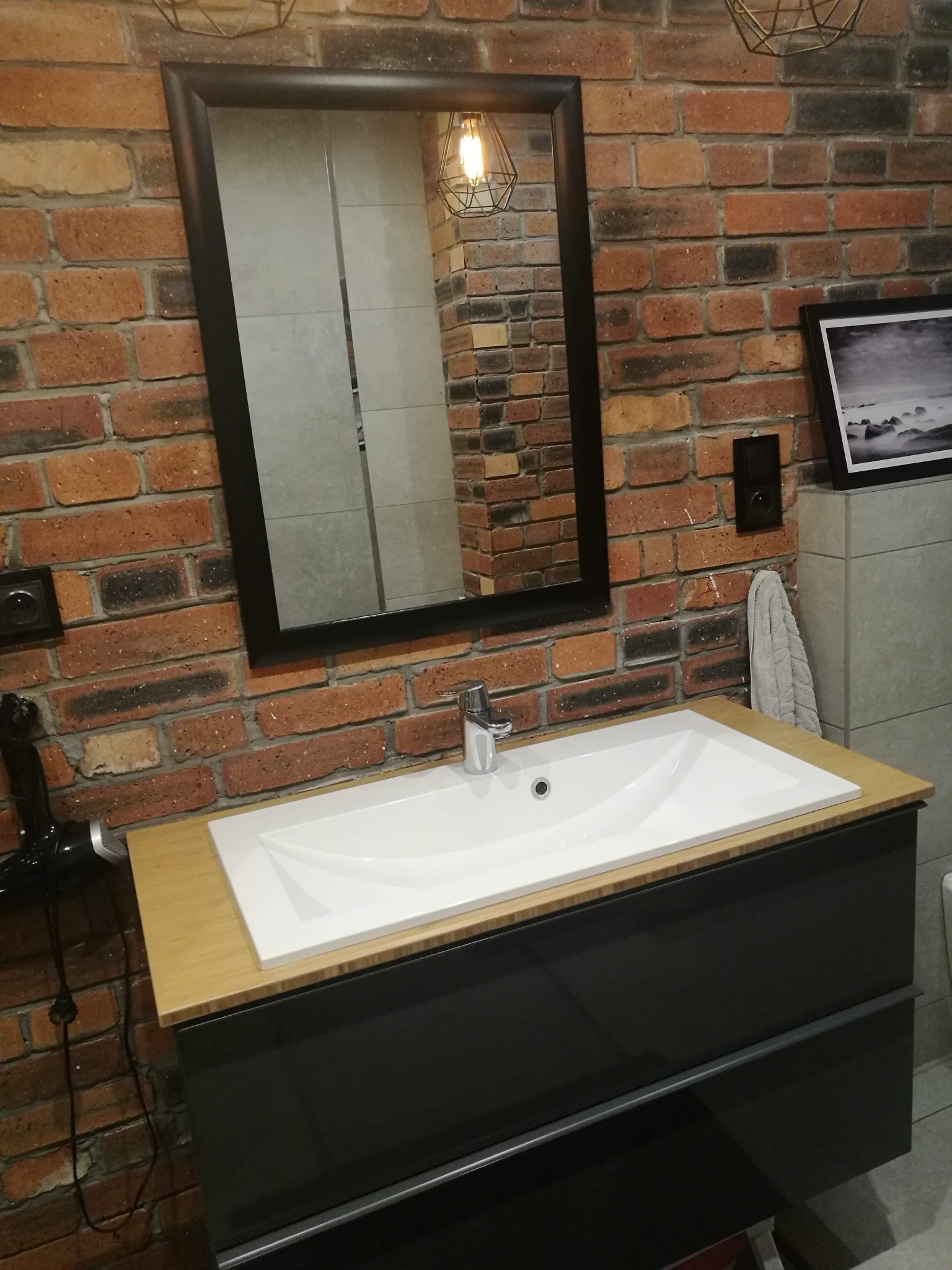 Nowoczesna łazienka z umywalką nablatową i szafką w kolorze grafitowym, na tle ściany z czerwonej cegły i lustra w czarnej ramie, widoczny czarny włącznik światła i suszarka do włosów.