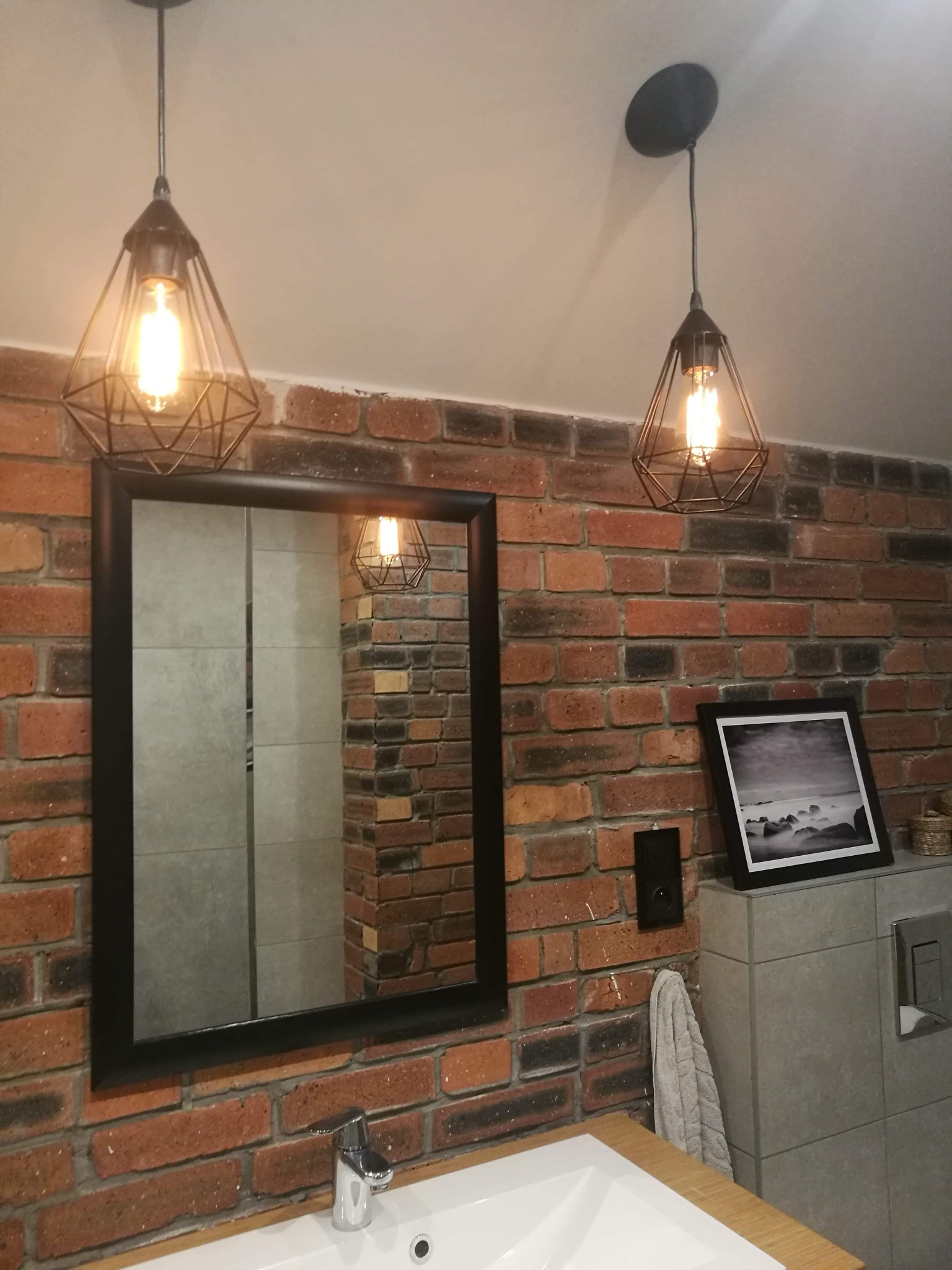 Ukończona łazienka z ceglaną ścianą, wiszącym lustrem w czarnej ramie, nowoczesną umywalką i dwoma geometrycznymi lampami wiszącymi z żarówkami Edisona.