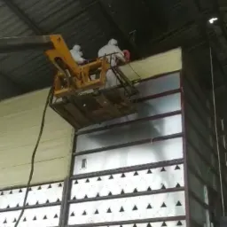 Pracownicy w kombinezonach ochronnych na podnośniku koszowym aplikują izolację na ścianę hali przemysłowej.