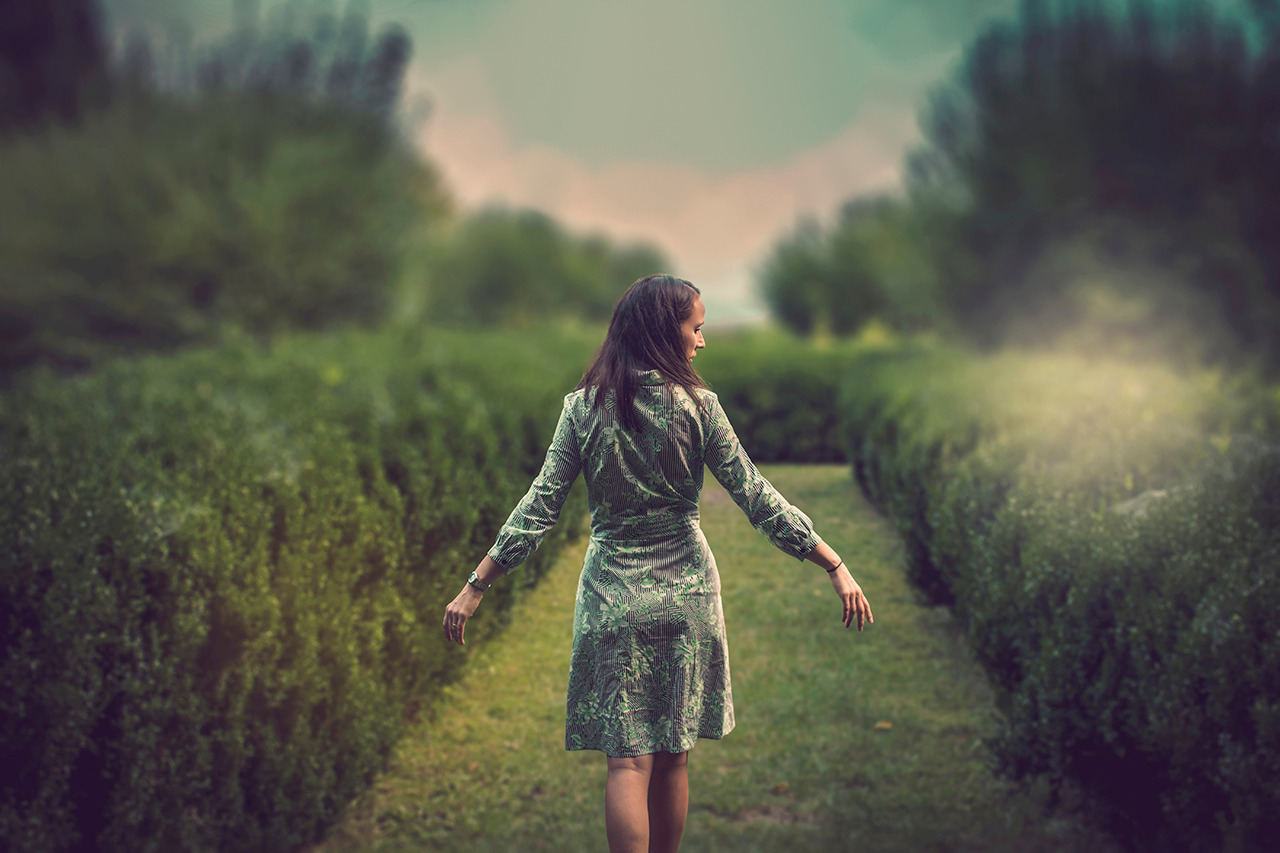 Kobieta w zielonej sukience z motywem liści spaceruje ścieżką wytyczoną przez równo przycięte żywopłoty, z rozłożonymi ramionami, w kierunku rozmytego tła z drzewami.