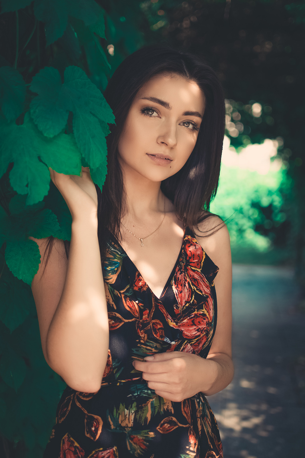 Portret młodej kobiety z długimi, ciemnymi włosami, w kwiecistej sukience, pozującej na tle zielonych liści i rozmytego tła alejki.