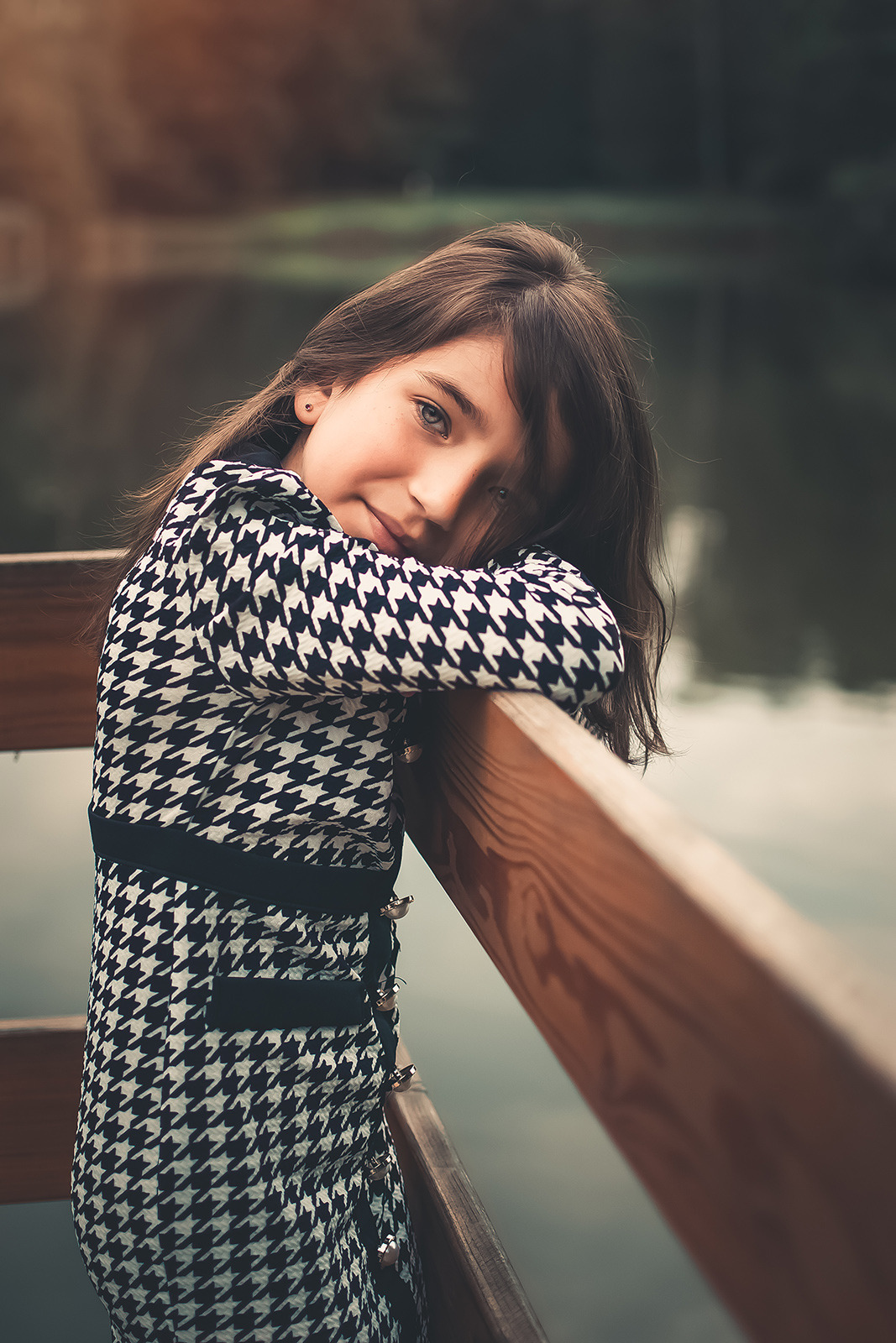 Portret dziewczynki w sukience w pepitkę, opierającej się o drewnianą barierkę z widokiem na jezioro w tle.