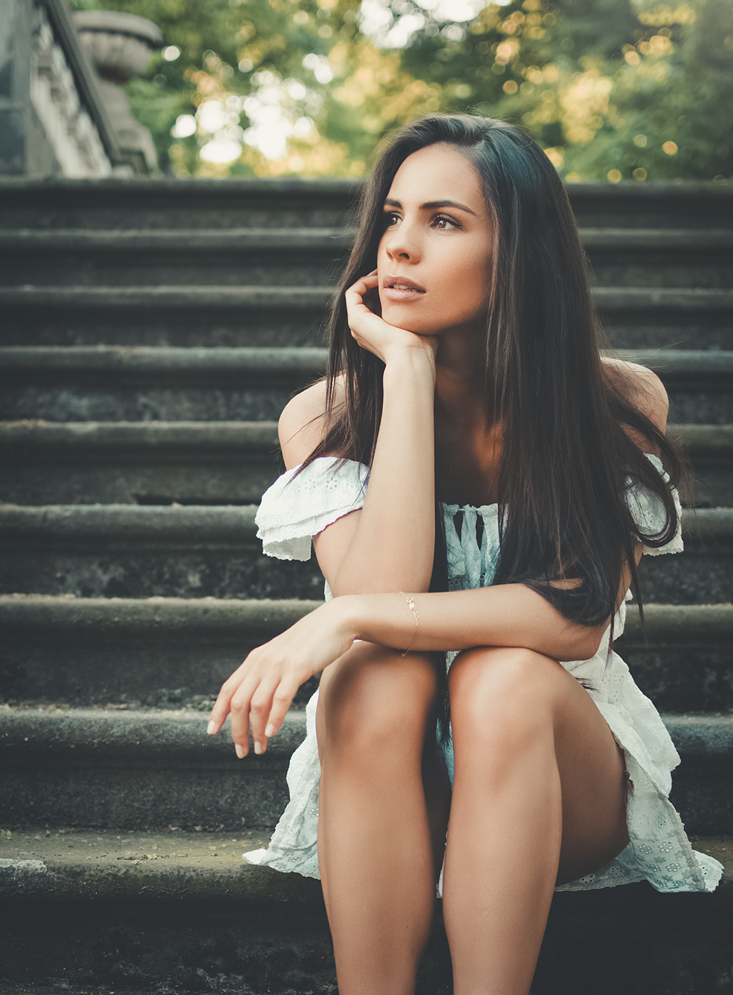 Zamyślona brunetka w białej sukience siedzi na kamiennych schodach, podpierając brodę dłonią i spoglądając w górę.