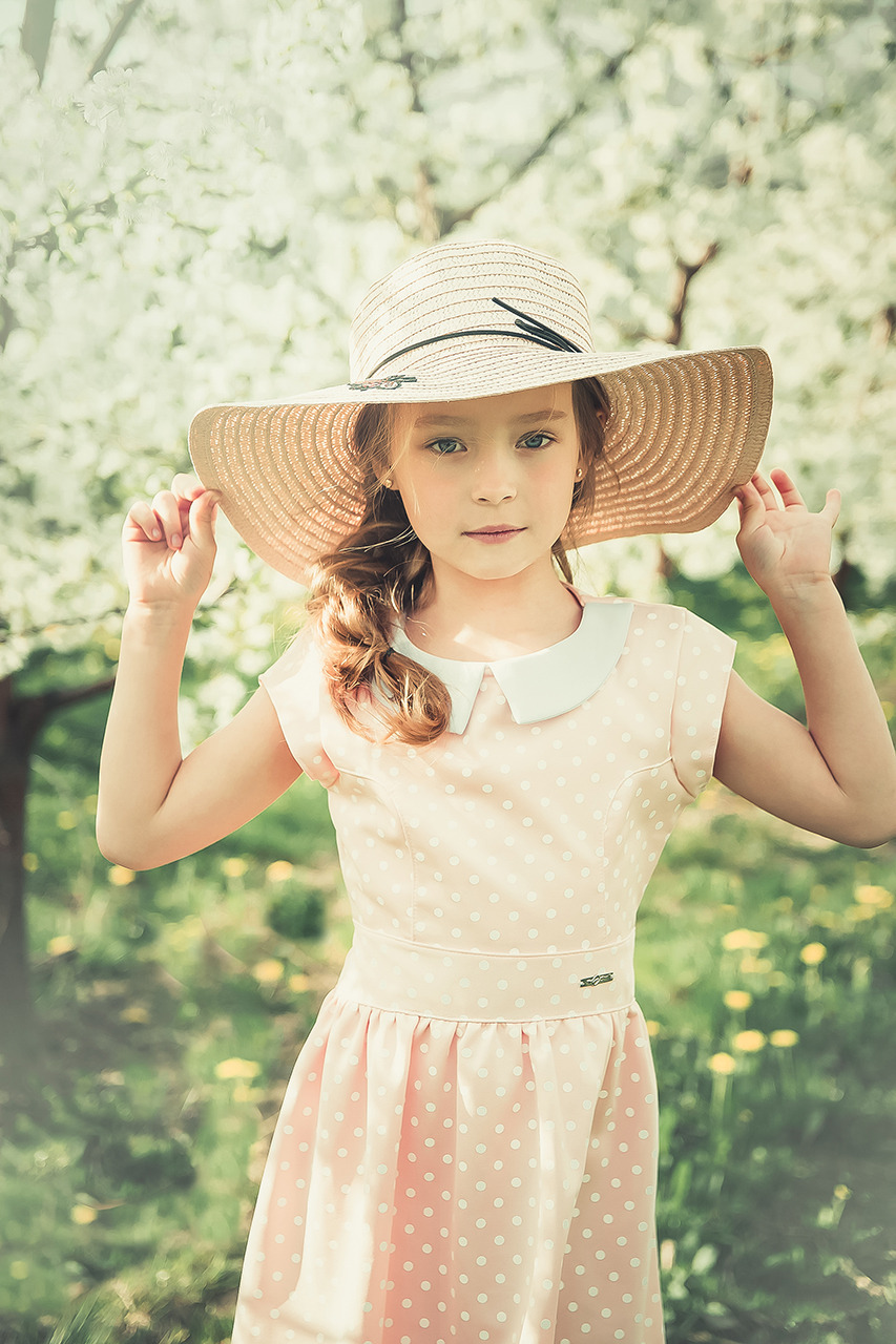 Portret dziewczynki w słomkowym kapeluszu i sukience w kropki na tle kwitnących drzew owocowych.