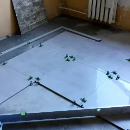 Układanie jasnoszarych płytek podłogowych z użyciem poziomicy i klipsów poziomujących w trakcie prac remontowych. W tle widoczny grzejnik i surowe ściany.