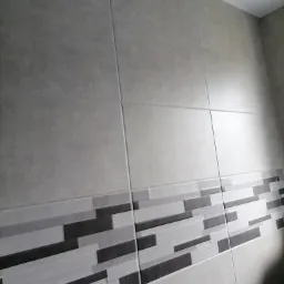 Wykończona ściana łazienki z szarymi płytkami, w dolnej części dekoracyjny pas z szarych i czarnych prostokątnych płytek.