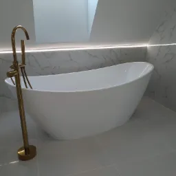 Nowoczesna łazienka z wolnostojącą białą wanną i złotą baterią podłogową, marmurowe płytki na ścianach, oświetlenie LED pod sufitem.