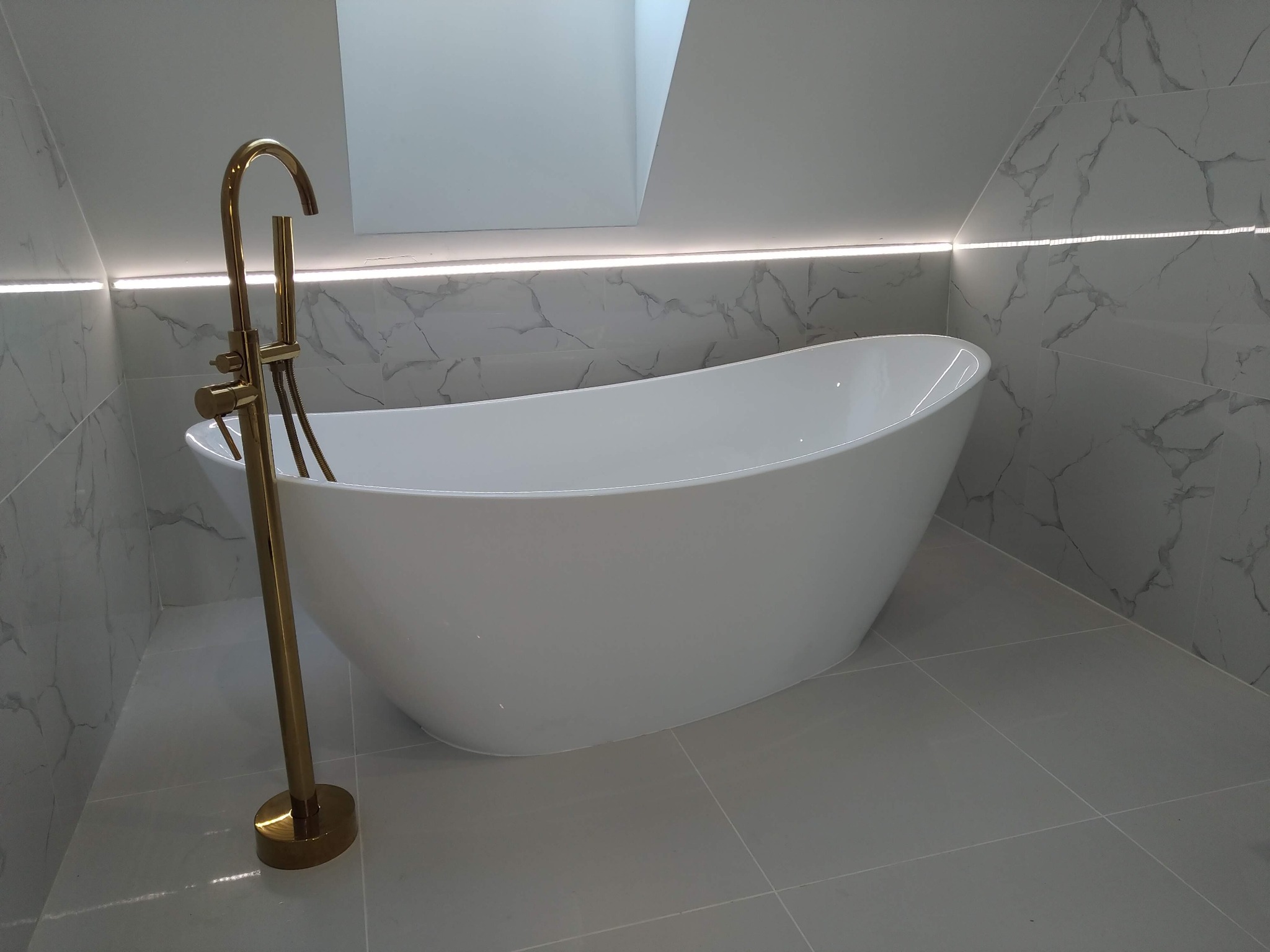 Nowoczesna łazienka z wolnostojącą białą wanną i złotą baterią podłogową, marmurowe płytki na ścianach, oświetlenie LED pod sufitem.