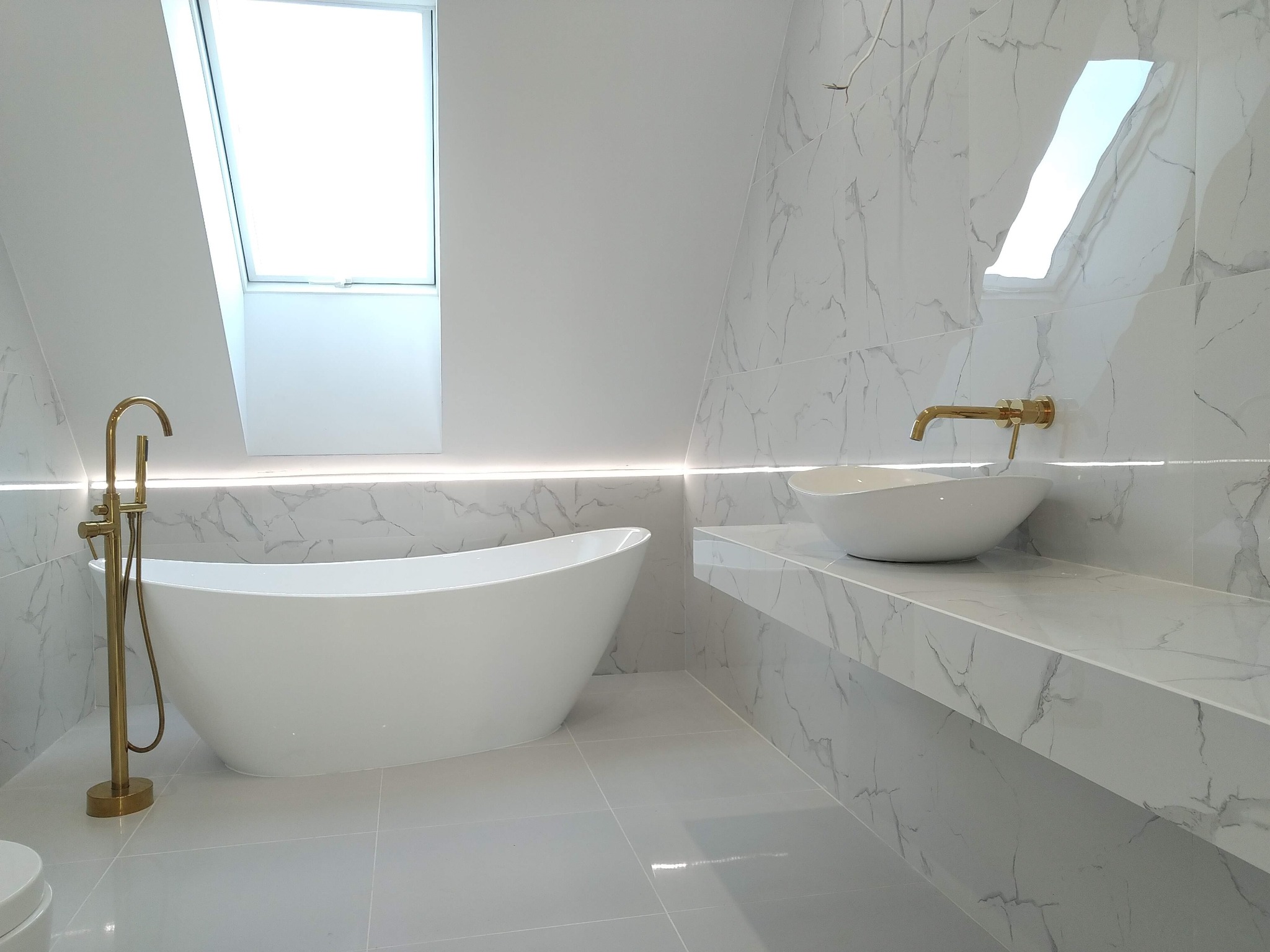 Nowoczesna łazienka w bieli i szarości z marmurkowymi płytkami, wolnostojąca wanna z złotą armaturą i umywalka nablatowa pod oknem dachowym.
