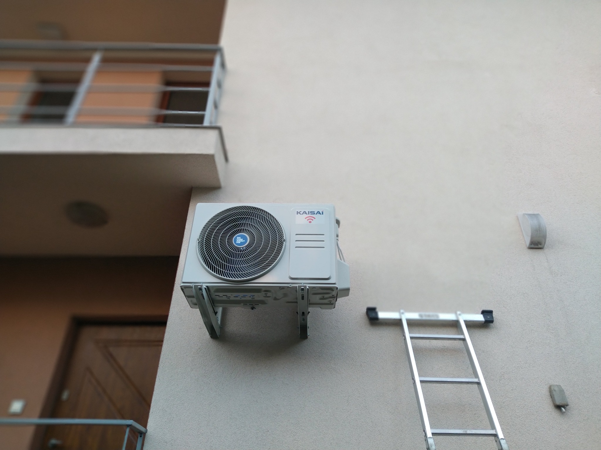 Zewnętrzna jednostka klimatyzatora marki Kaisai zamontowana na ścianie budynku, widoczna drabina aluminiowa oparta o ścianę obok urządzenia.